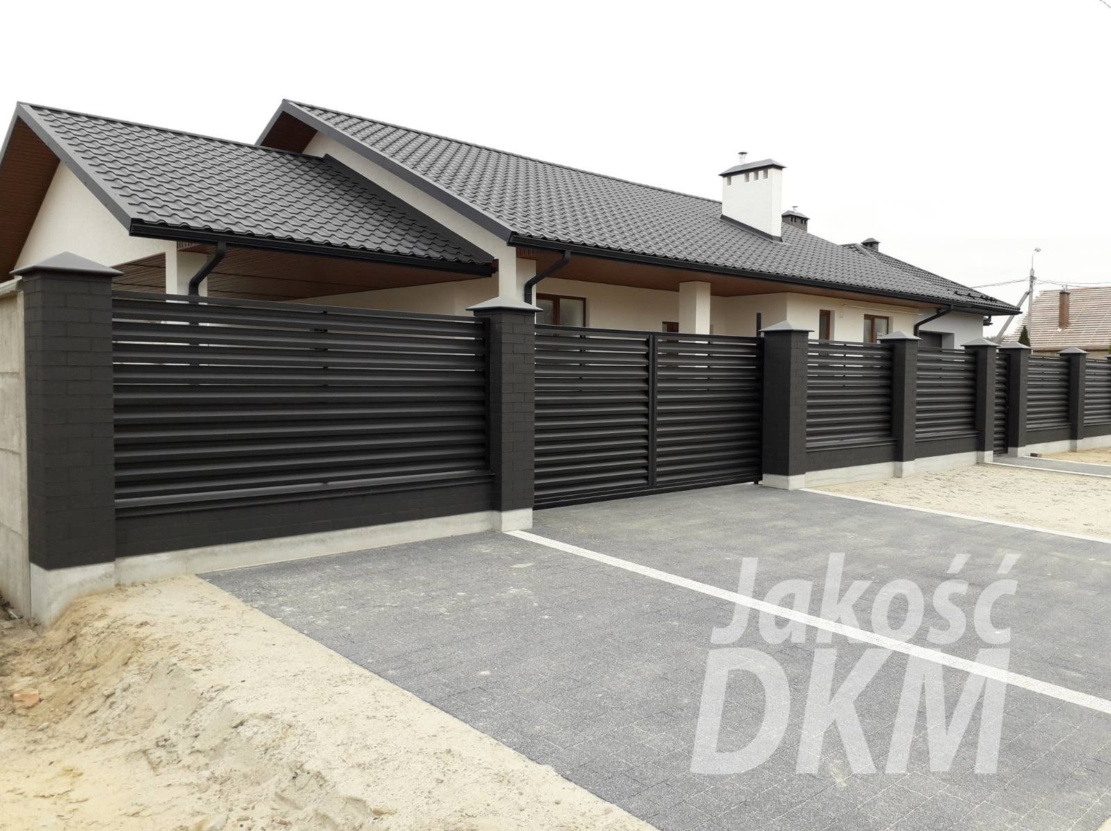 Nowoczesne, poziome ogrodzenie panelowe w kolorze antracytowym, z bramą przesuwną i słupkami z cegły klinkierowej, na tle domu z ciemną dachówką i kostki brukowej.
