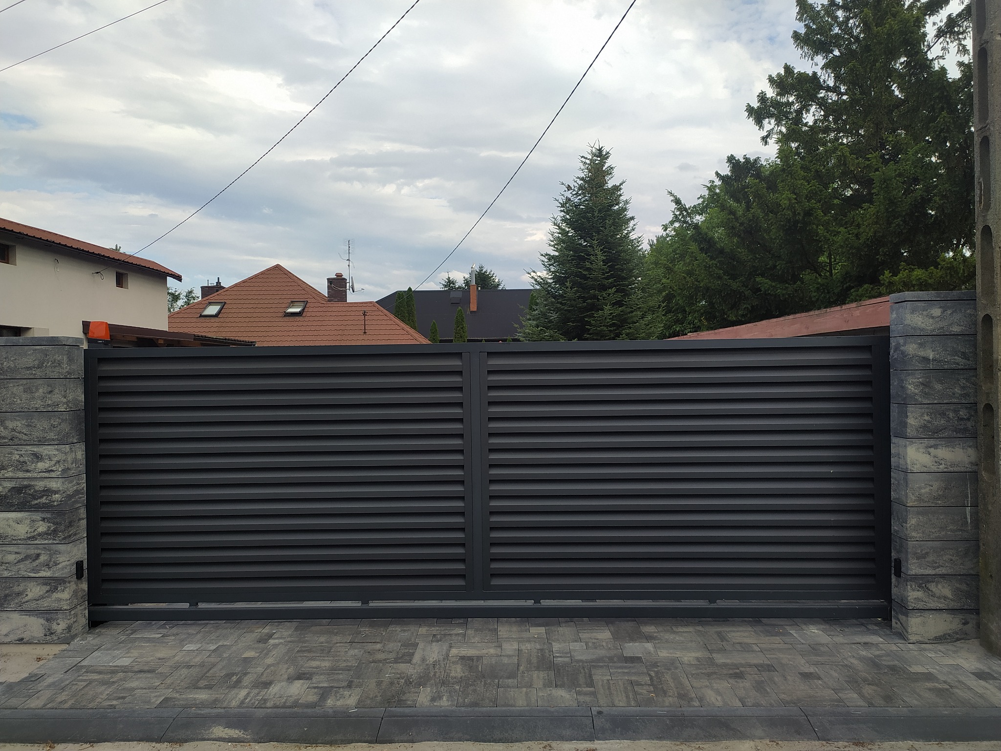 Nowoczesna przesuwna brama wjazdowa z poziomych profili, w kolorze grafitowym, zamontowana na słupkach z szarej kostki łupanej, z widocznym podjazdem z kostki brukowej.