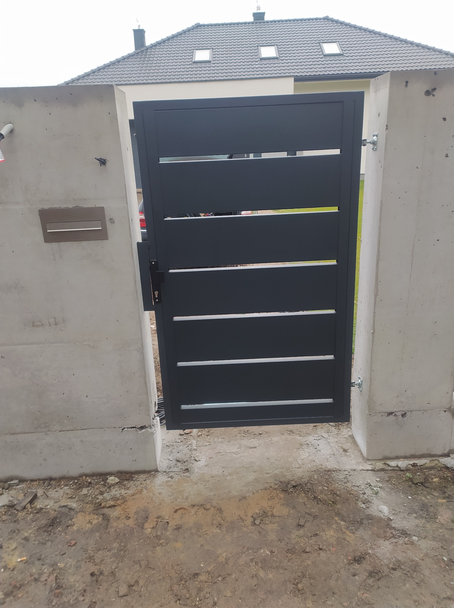 Nowoczesna, grafitowa furtka z poziomymi szczeblami, zamontowana między betonowymi słupkami z widoczną skrzynką na listy, na tle domu z szarą dachówką.