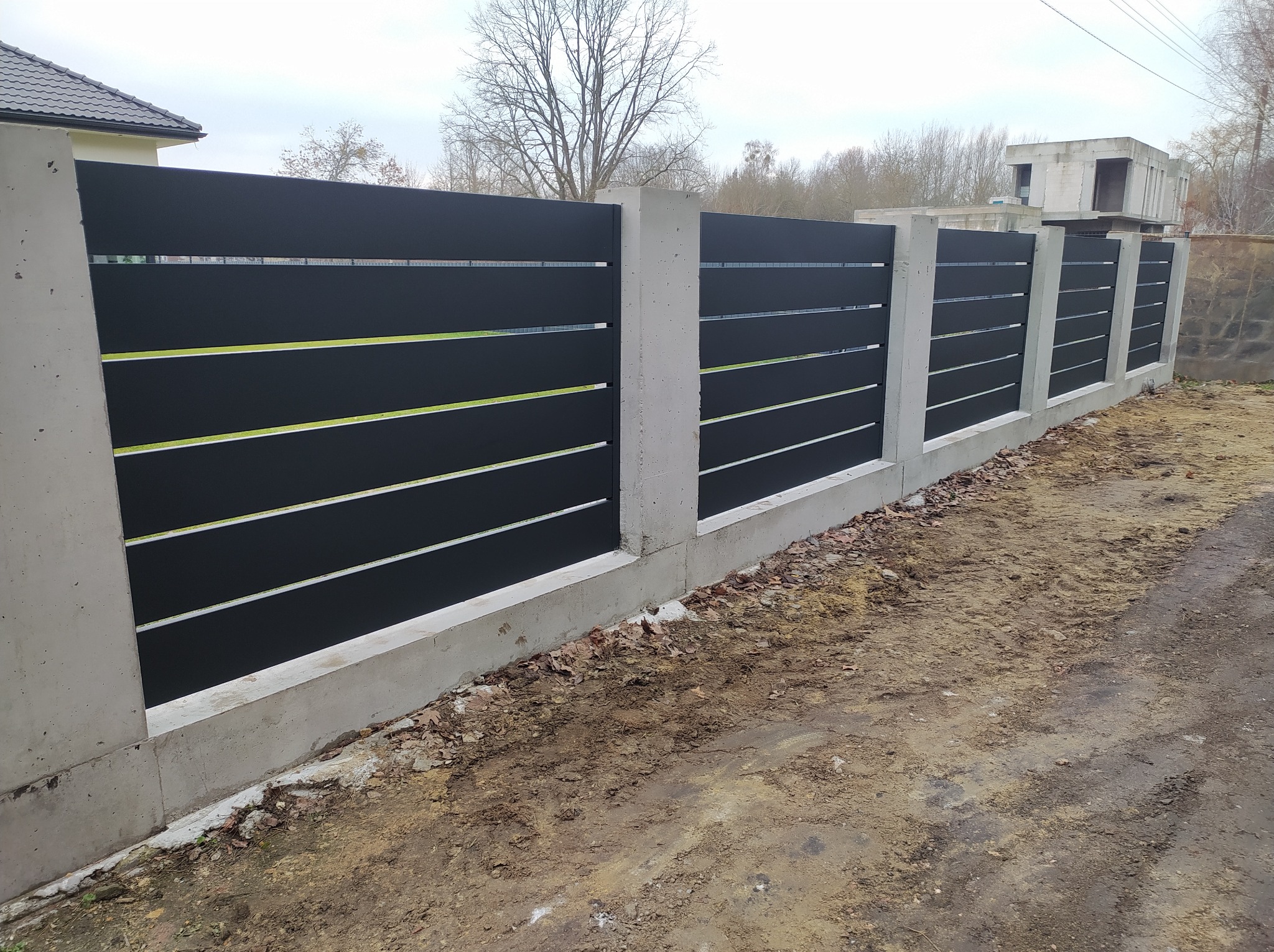 Nowoczesne ogrodzenie z czarnych paneli z poziomymi szczelinami, osadzone w betonowych słupkach i fundamencie, na tle jesiennego krajobrazu z widocznym niedokończonym budynkiem.