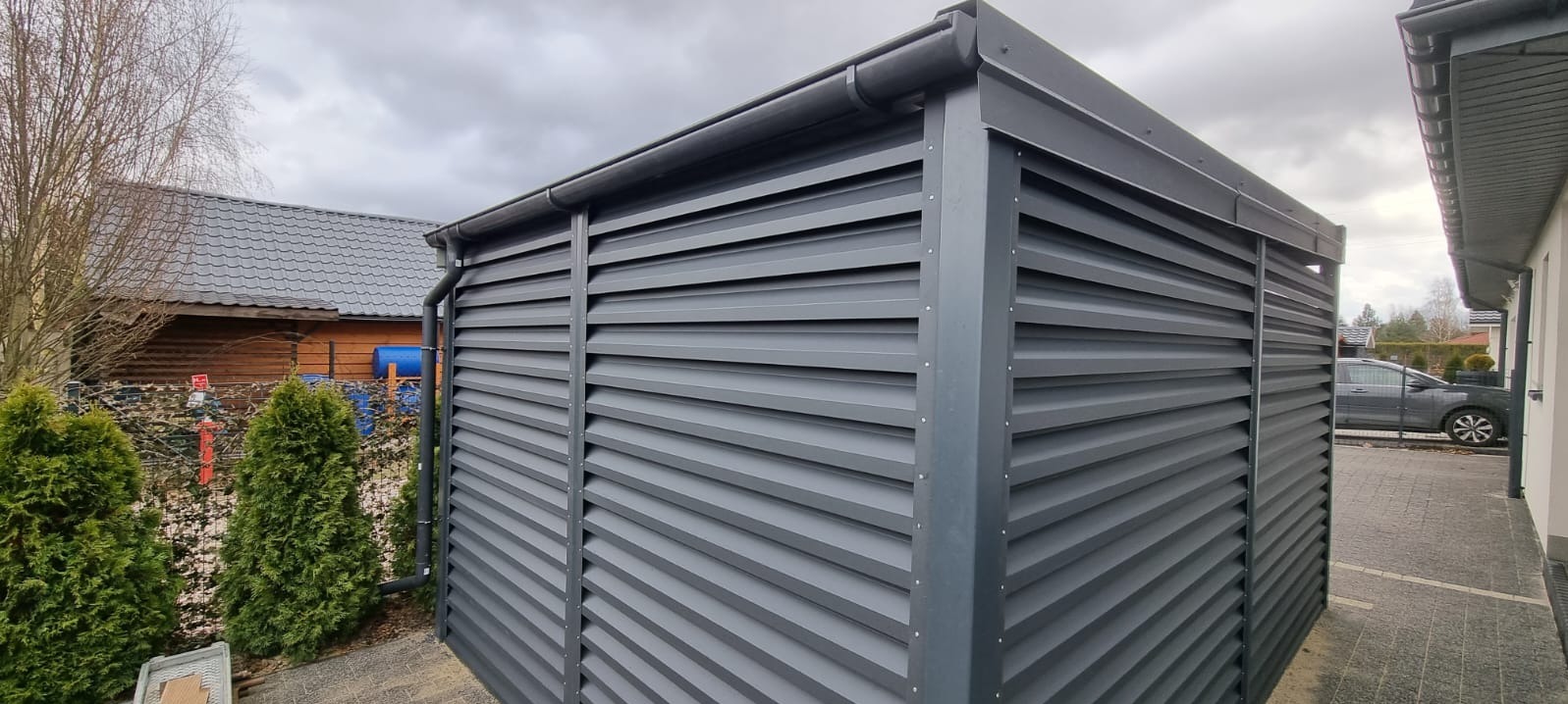 Nowoczesna, szara konstrukcja z metalowych żaluzji, prawdopodobnie osłona śmietnika lub element ogrodzenia posesji, z widocznym systemem odprowadzania wody deszczowej i fragmentem domu w tle.