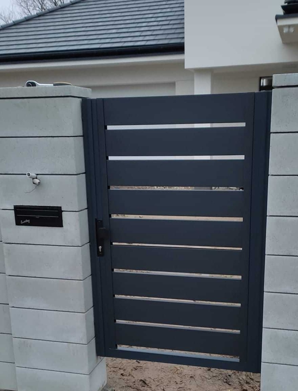 Nowoczesna brama z poziomymi szczeblami, wbudowana w murowany słupek z szarej kostki, skrzynka na listy po lewej stronie, tło stanowi elewacja domu z ciemnym dachem.