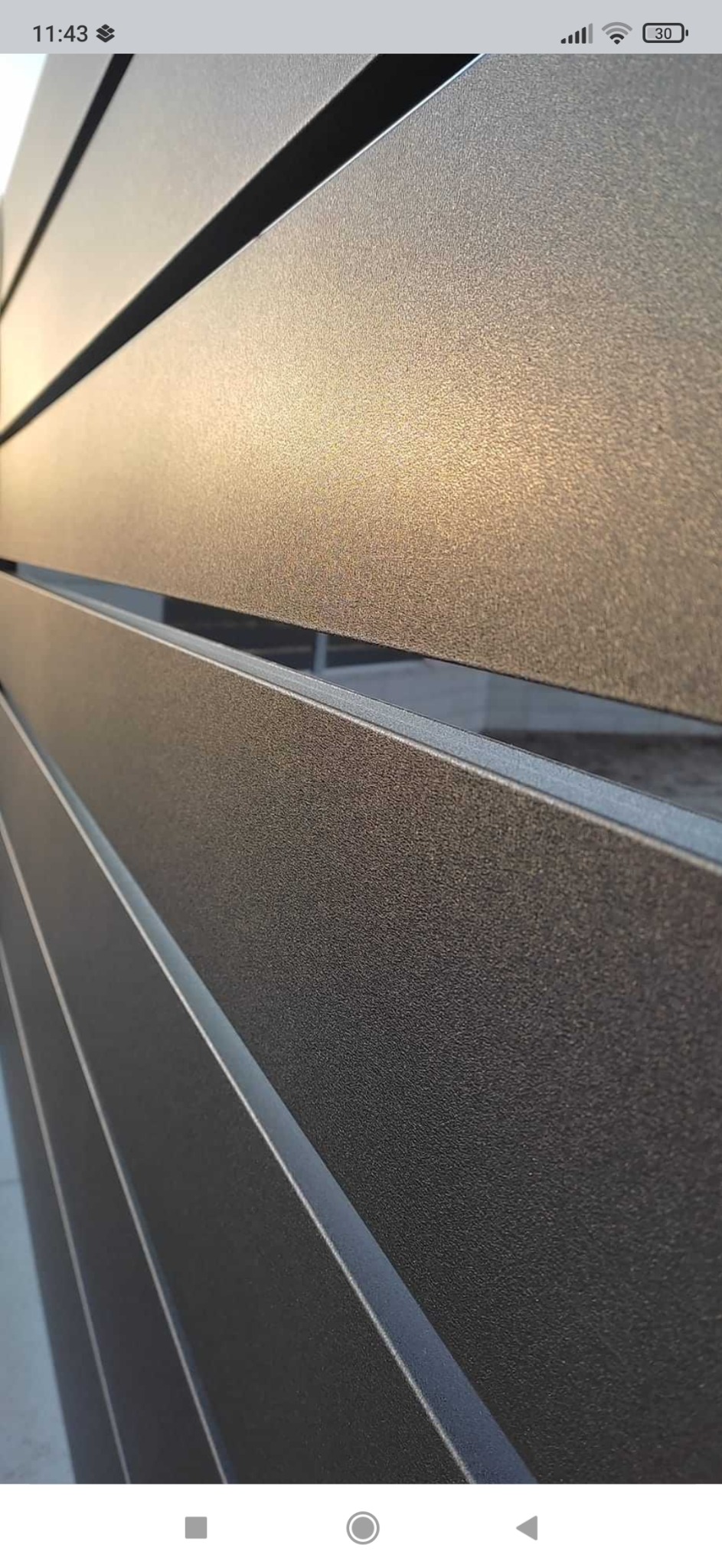 Fragment nowoczesnego, poziomego ogrodzenia z metalowych profili o szarej, teksturowanej powierzchni, widoczny z bliska pod kątem.