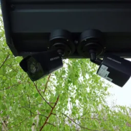 Dwie czarne kamery monitoringu Hikvision zamontowane pod zadaszeniem, widoczne z dołu na tle zielonych liści drzew.