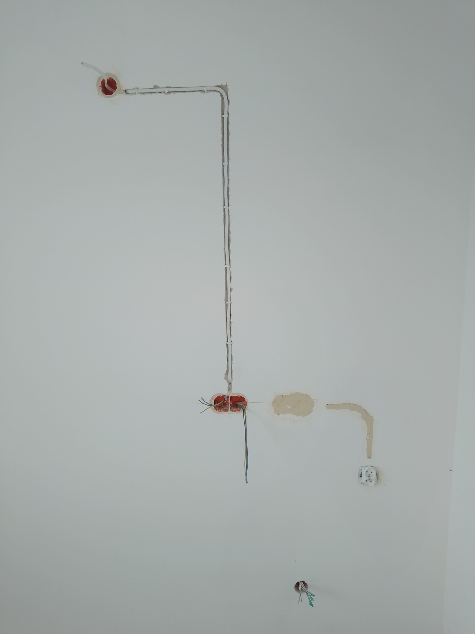 Biała ściana z widocznymi elementami instalacji elektrycznej: puszkami, przewodami w peszlach i gniazdkiem, po śladach na ścianie widać, że prace nie zostały jeszcze zakończone.