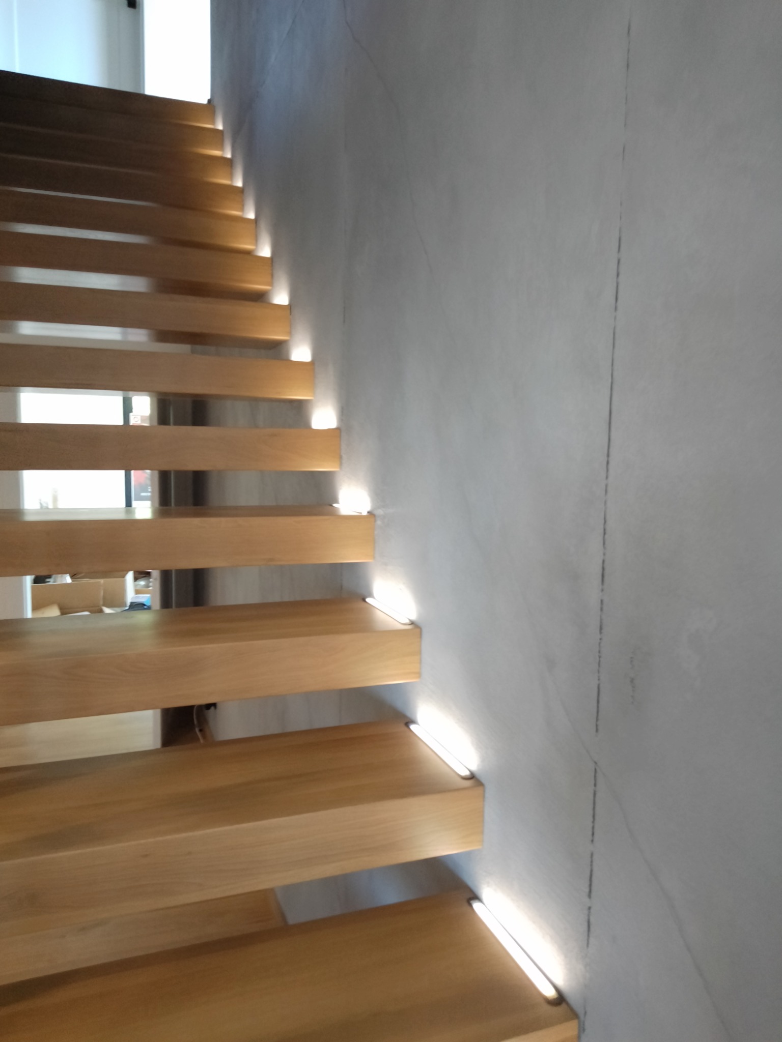 Drewniane schody z podświetleniem LED, zamontowanym pod każdym stopniem, przylegające do szarej, betonowej ściany. Widoczne okno w tle.