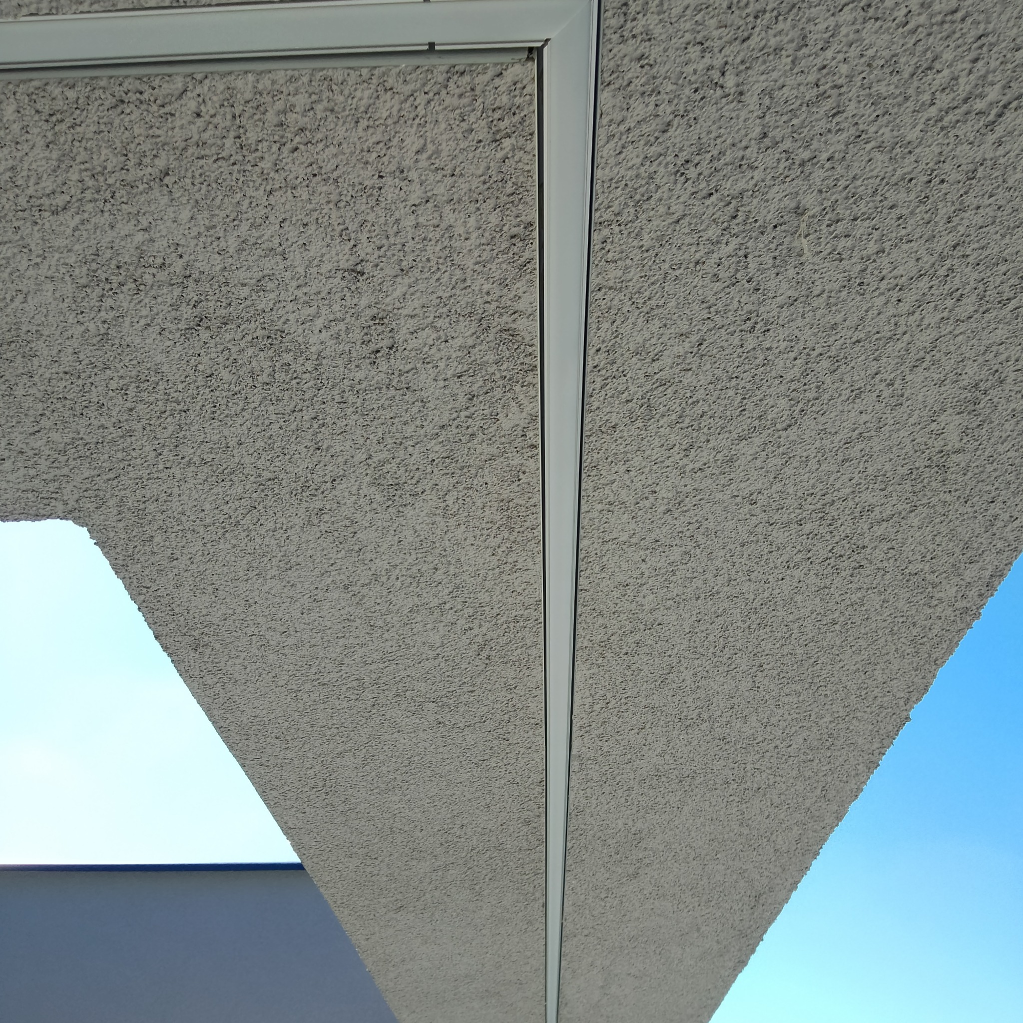 Szary, chropowaty sufit z widoczną białą listwą montażową i fragmentami błękitnego nieba w tle.