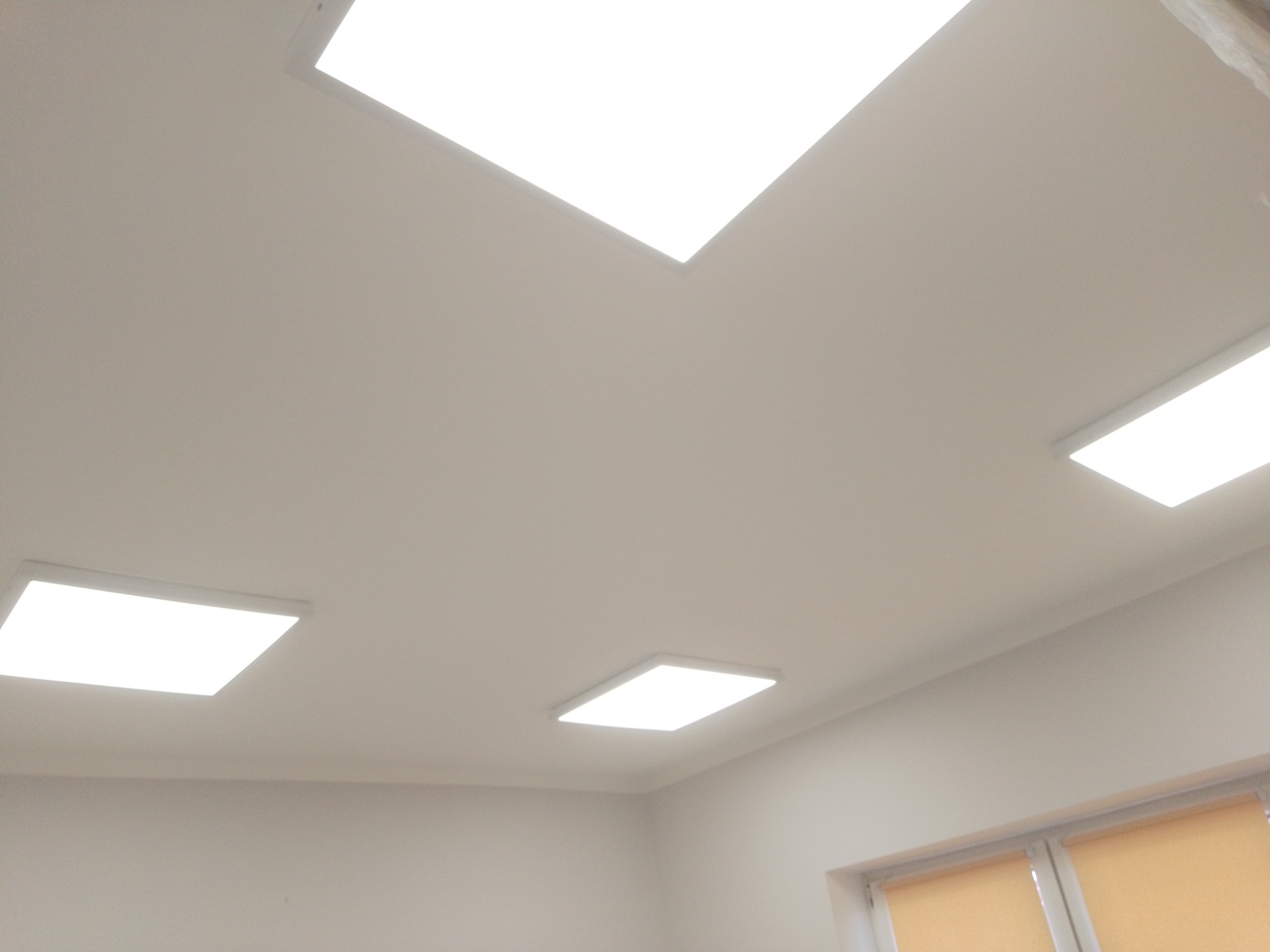 Biały sufit z czterema kwadratowymi panelami LED, widok pod kątem, okno z roletami w tle.