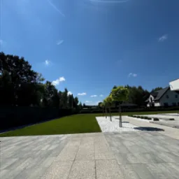 Widok na starannie zaprojektowany ogród z równo przystrzyżonym trawnikiem, białymi kamieniami ozdobnymi i rzędem kulistych drzewek na tle błękitnego nieba, z widocznymi elementami nowoczesnej...