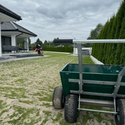 Usługi ogrodnicze Kamil Sosnowski - Zielony wózek z czterema kołami na trawniku posypanym piaskiem, w tle nowoczesny dom z płaskim dachem i osoba siedząca na schodach.