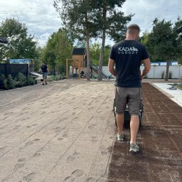 Usługi ogrodnicze Kamil Sosnowski - Pracownik w koszulce z logo firmy KADA Ogrody używa walca do wyrównywania powierzchni pod przyszły trawnik, widoczne ślady stóp na piasku, w tle drugi pracownik z grabiami oraz plac zabaw.