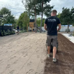 Pracownik w koszulce z logo firmy KADA Ogrody używa walca do wyrównywania powierzchni pod przyszły trawnik, widoczne ślady stóp na piasku, w tle drugi pracownik z grabiami oraz plac zabaw.