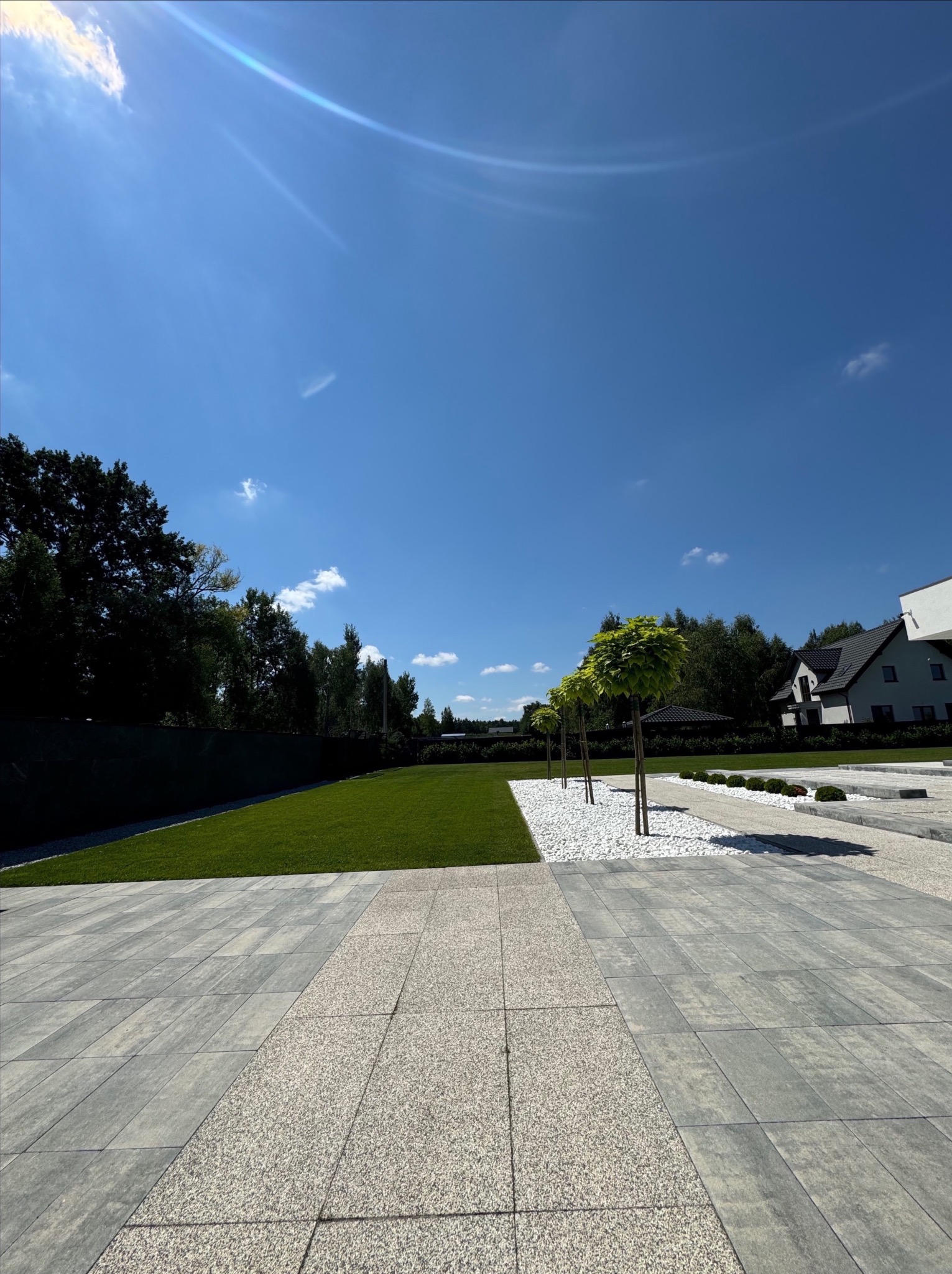 Widok na starannie zaprojektowany ogród z równo przystrzyżonym trawnikiem, białymi kamieniami ozdobnymi i rzędem kulistych drzewek na tle błękitnego nieba, z widocznymi elementami nowoczesnej...