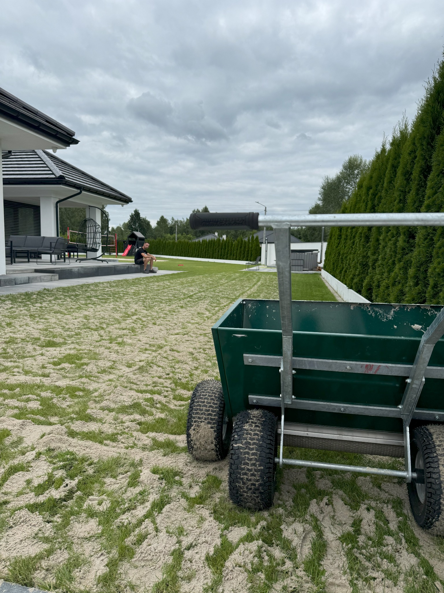 Zielony wózek z czterema kołami na trawniku posypanym piaskiem, w tle nowoczesny dom z płaskim dachem i osoba siedząca na schodach.