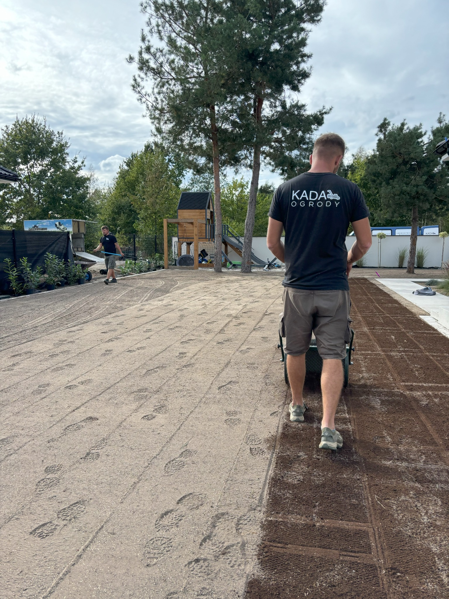 Pracownik w koszulce z logo firmy KADA Ogrody używa walca do wyrównywania powierzchni pod przyszły trawnik, widoczne ślady stóp na piasku, w tle drugi pracownik z grabiami oraz plac zabaw.