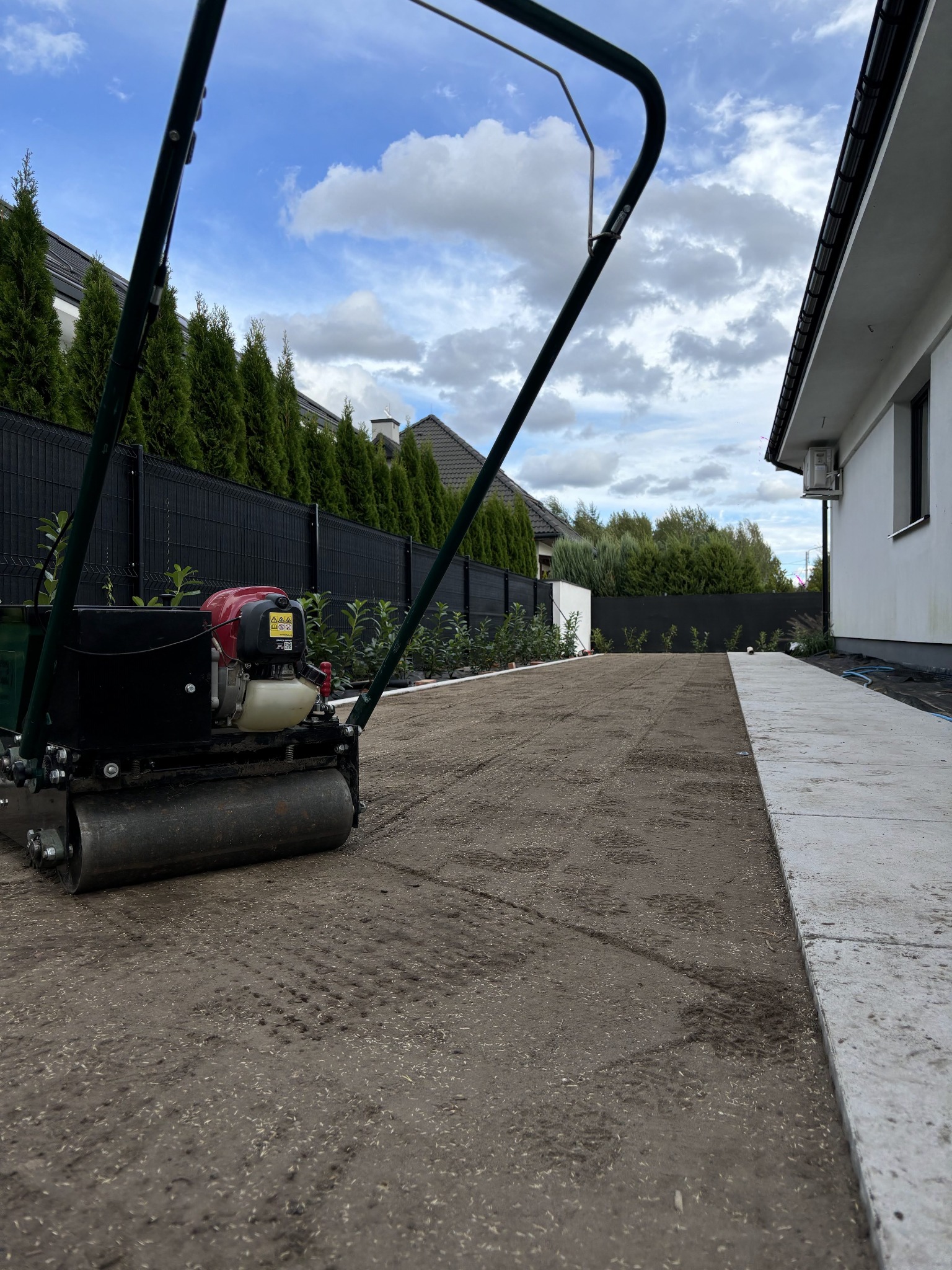 Walec ogrodowy zagęszcza glebę przygotowaną pod trawnik w przydomowym ogrodzie; widoczne ślady walca na ziemi, świeżo posiana trawa, obrzeże z kostki i budynek w tle.