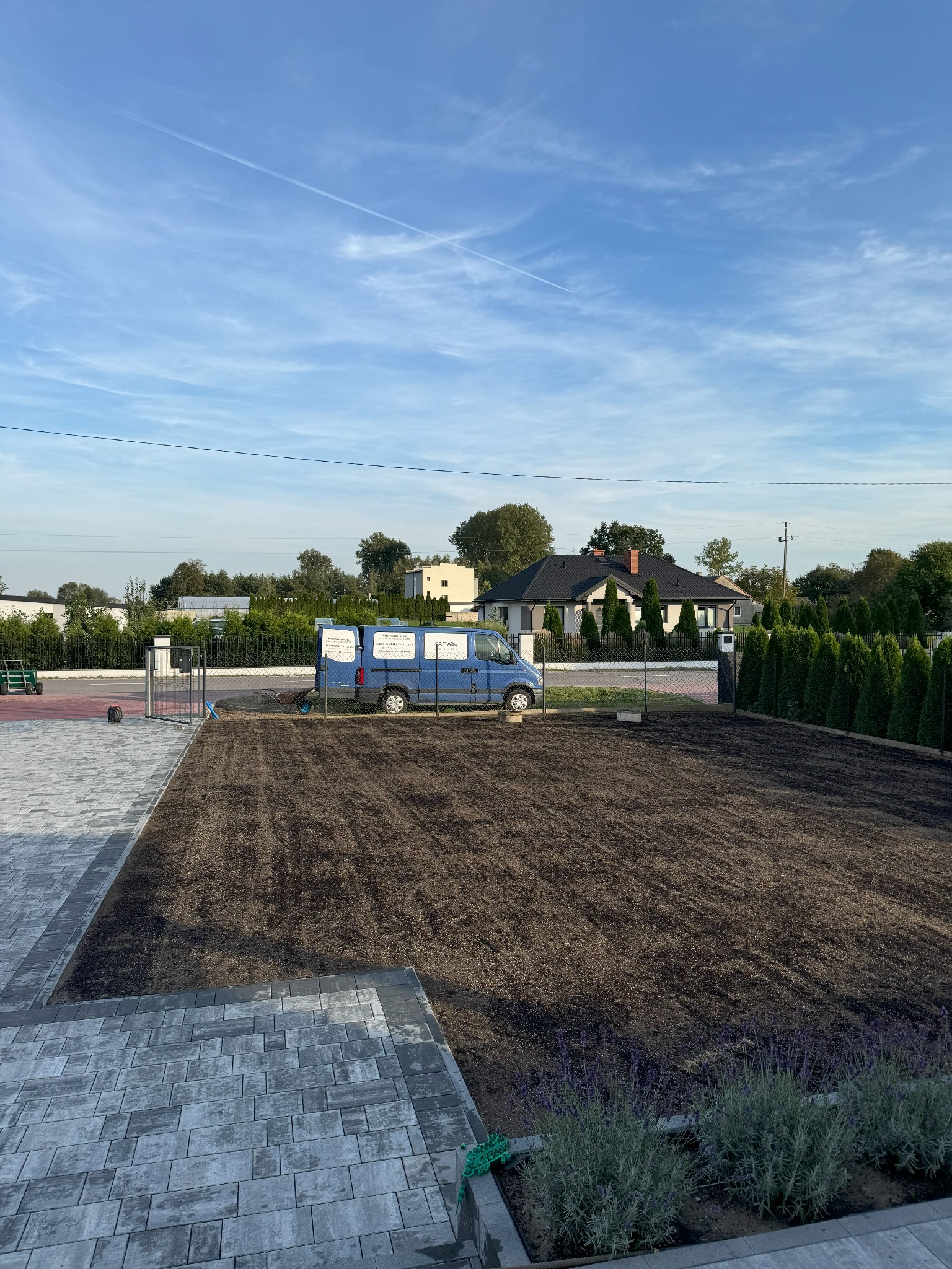 Przygotowany teren pod trawnik z widoczną ziemią, obok kostka brukowa i zaparkowany niebieski bus firmy ogrodniczej na tle domu jednorodzinnego i błękitnego nieba.