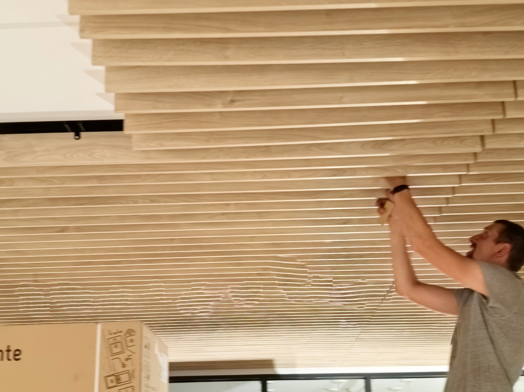 Montaż drewnianych paneli na suficie przez mężczyznę, widoczny fragment kartonu z instrukcjami, oświetlenie sufitowe w tle.