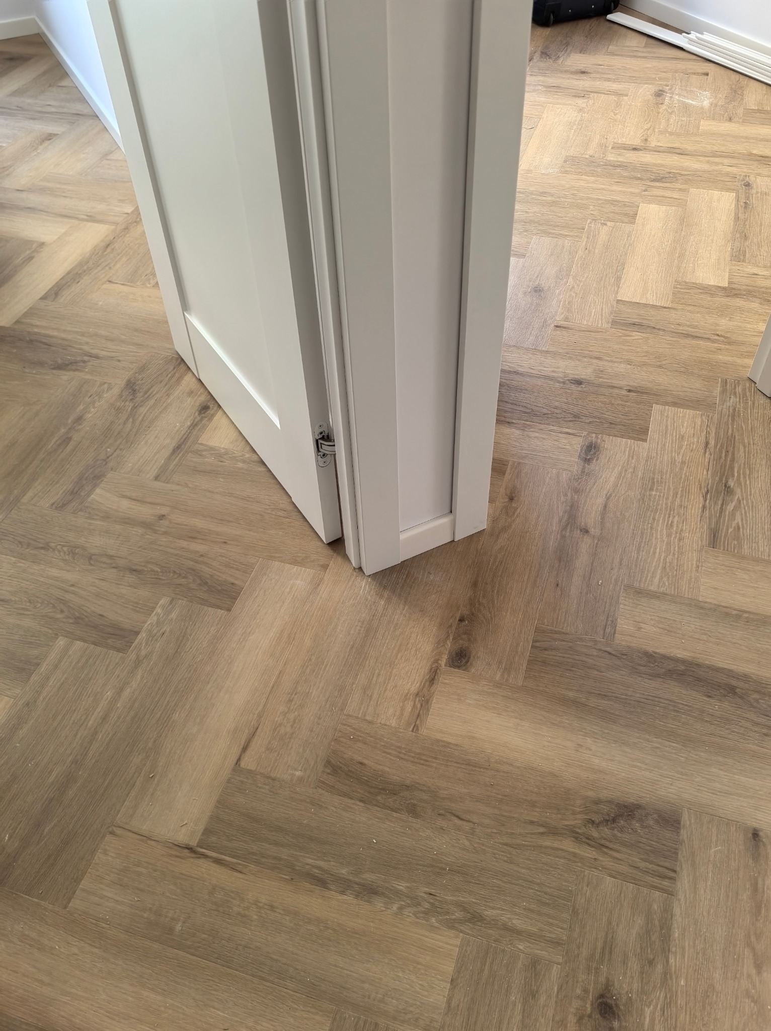 Ułożone panele podłogowe w jodełkę francuską, widoczne przez otwarte, białe drzwi z zawiasami.