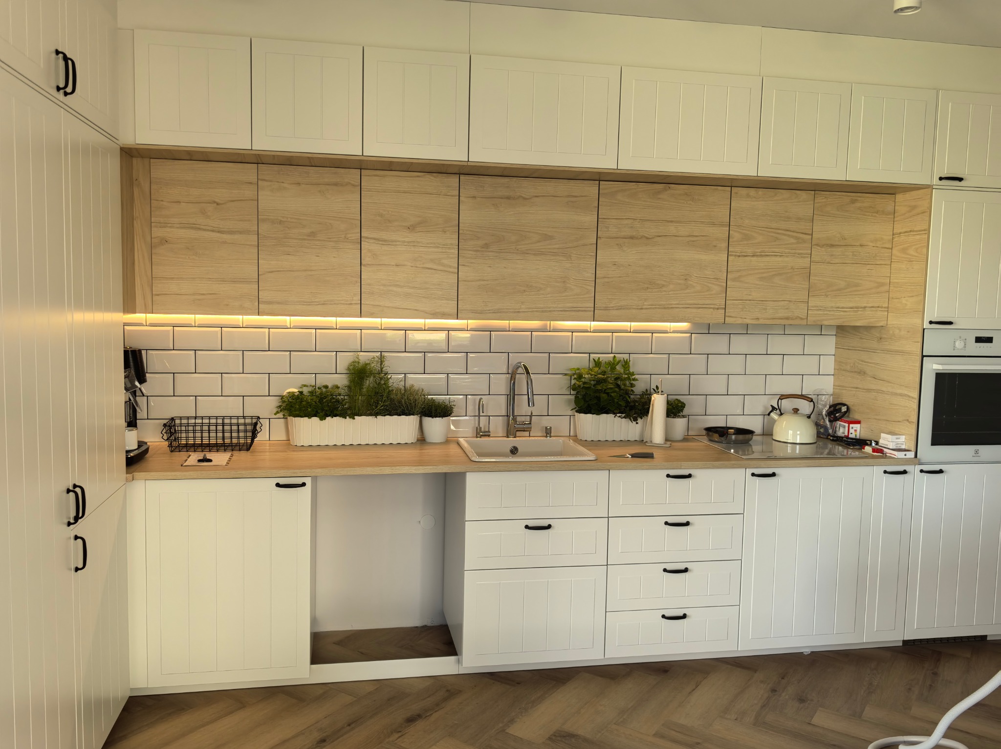 Jasna kuchnia z białymi szafkami z frezowanymi frontami i drewnopodobnymi górnymi szafkami, białe płytki metra na ścianie, zioła w doniczkach na blacie, podświetlenie LED pod szafkami.