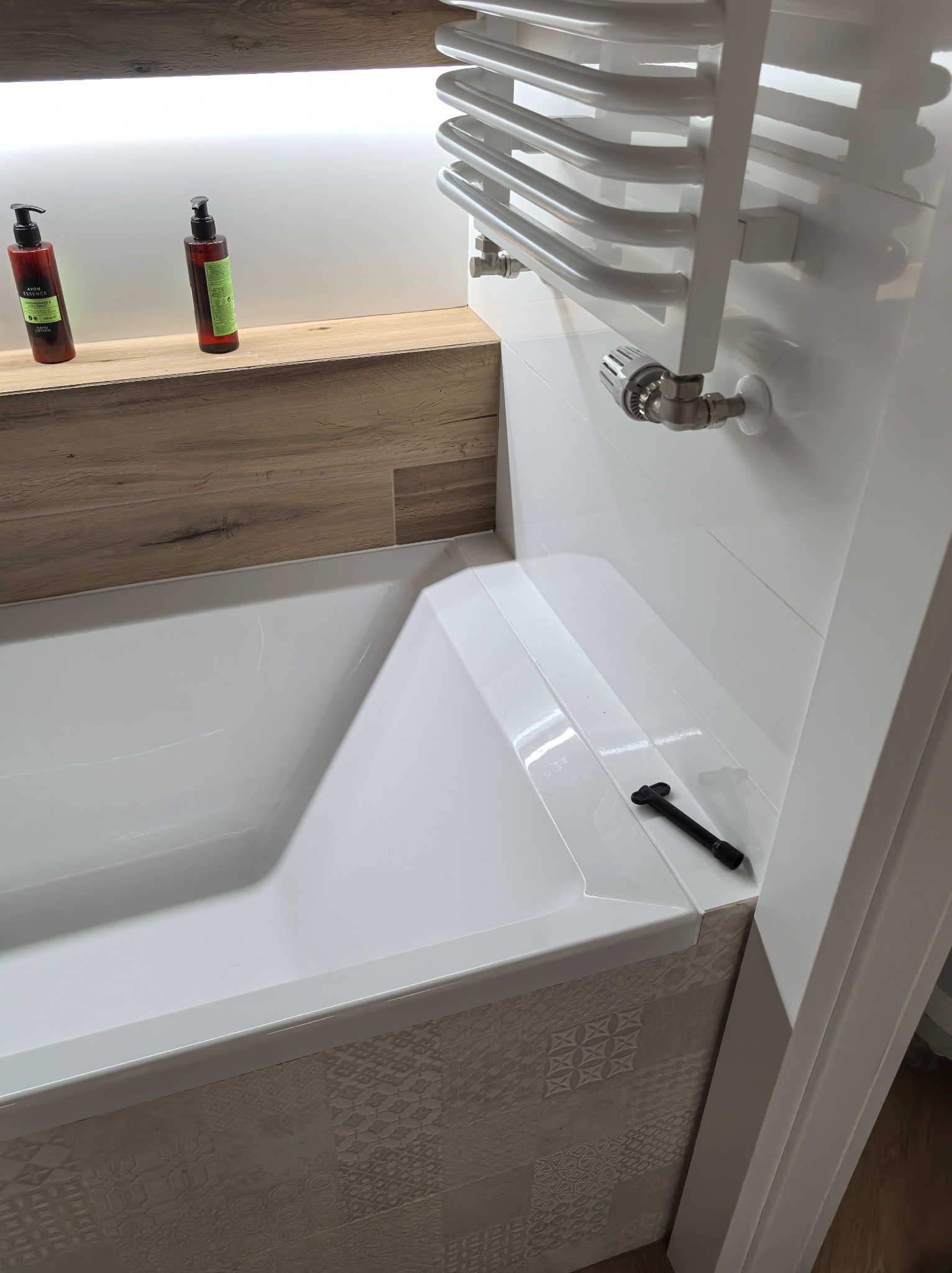 Biała wanna z grzejnikiem drabinkowym na ścianie z białych płytek, półka z drewnopodobną okleiną i butelkami kosmetyków, dekoracyjne płytki na obudowie wanny, czarny klucz do regulacji odpływu...