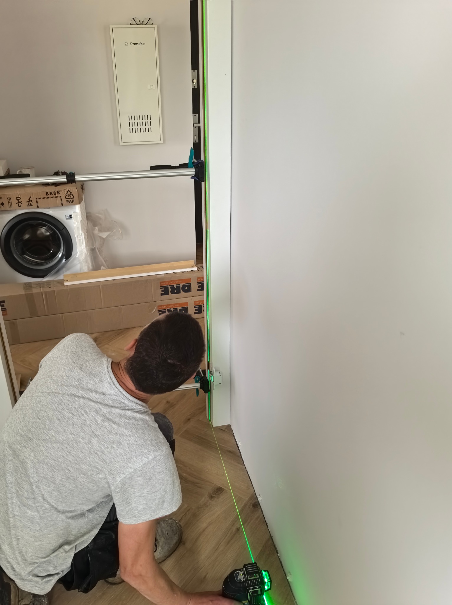 Instalacja ościeżnicy drzwiowej z użyciem poziomicy laserowej, widoczne kartony z ościeżnicami Dre, w tle pralka w folii. Ustawianie pionu ościeżnicy.