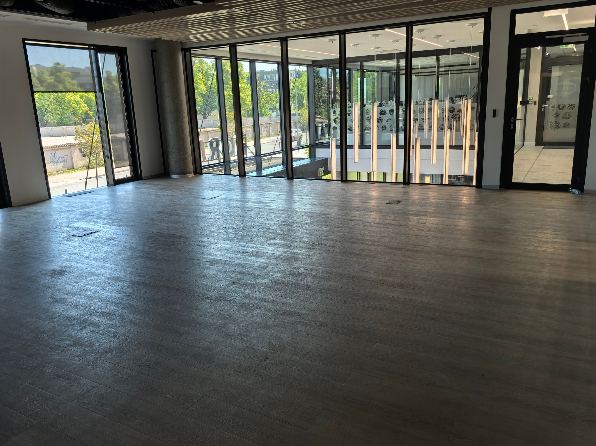 Wnętrze biura z parkietem ułożonym w deski, dużymi oknami z widokiem na zieleń i nowoczesnym oświetleniem wiszącym.