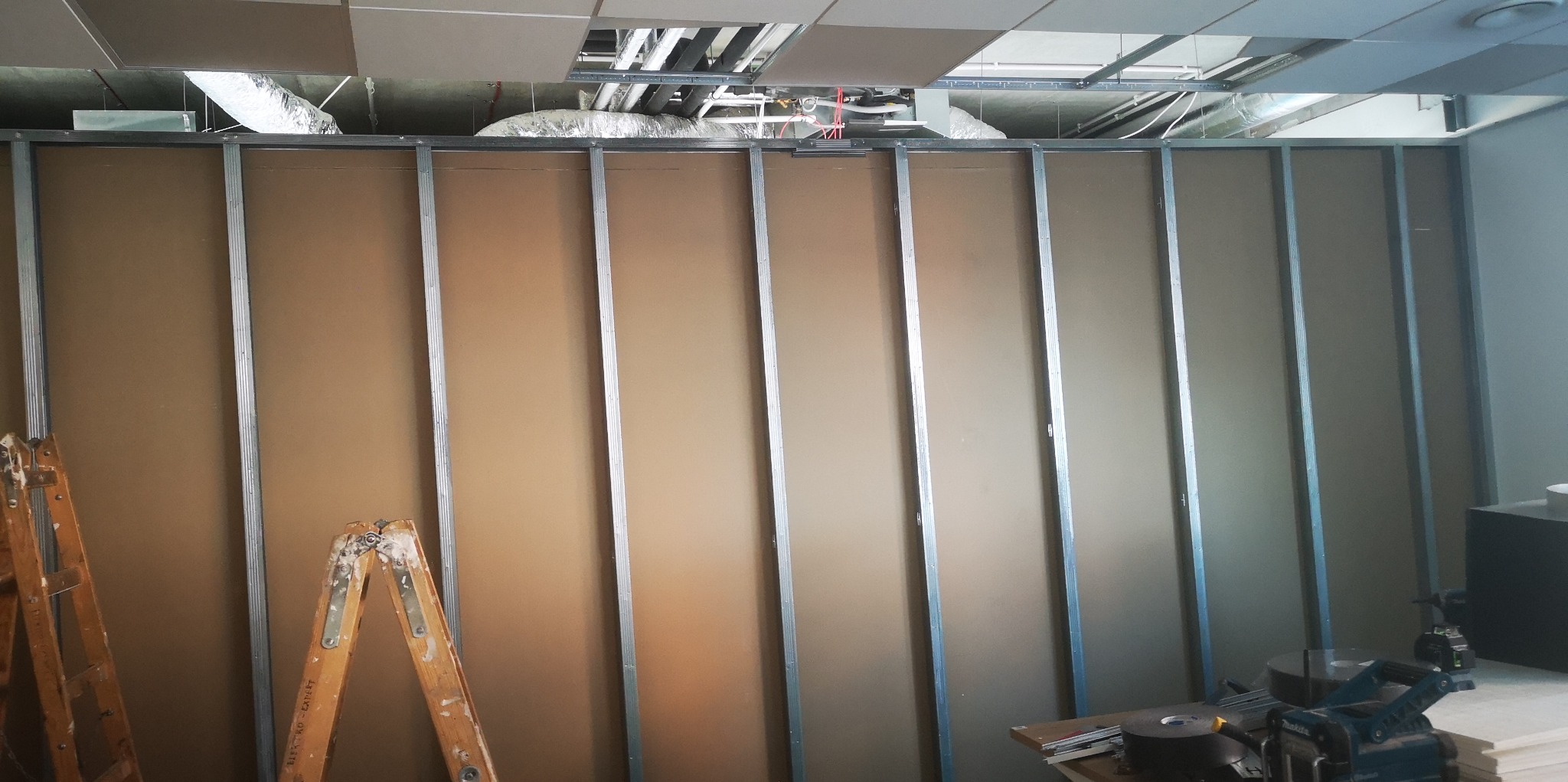 Montaż ścian działowych w biurze, widoczne płyty gipsowo-kartonowe na metalowym stelażu, dwie drewniane drabiny malarskie po lewej stronie, sufit podwieszany z widoczną instalacją wentylacyjną.