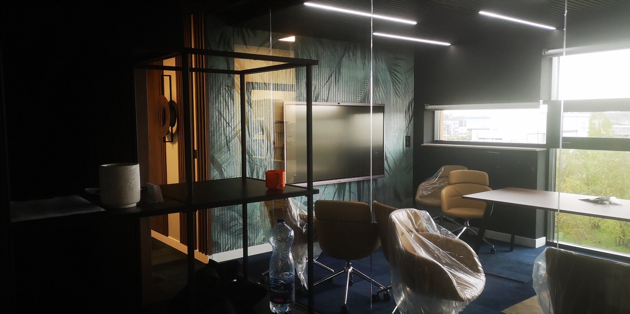 Biuro w trakcie aranżacji: krzesła zabezpieczone folią, widoczna butelka wody, ściana z motywem roślinnym i szklana ścianka działowa.