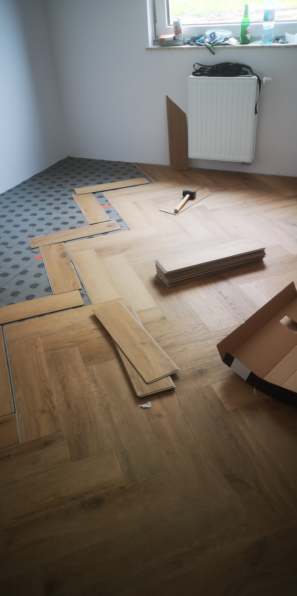 Fragment podłogi w trakcie montażu paneli w jodełkę; widoczny podkład, ułożone fragmenty oraz narzędzia i opakowanie.