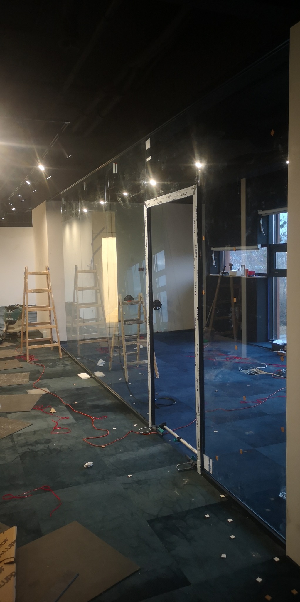 Montaż szklanej ścianki działowej w biurze, widoczne drabiny, poziomica laserowa i kable na podłodze z ciemną wykładziną.