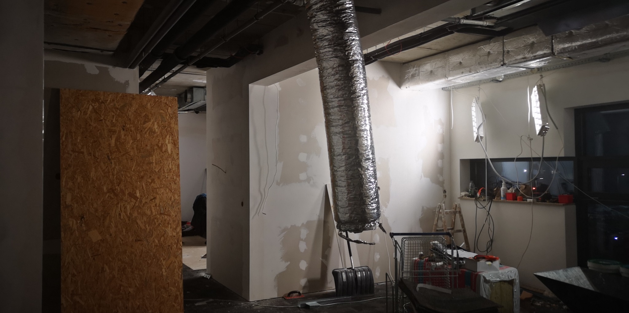 Wnętrze w trakcie prac przygotowawczych do wykończenia ścian, widoczne fragmenty płyty OSB, ściany ze śladami szpachlowania, aluminiowy przewód wentylacyjny, prowizoryczne oświetlenie, narzędzia...