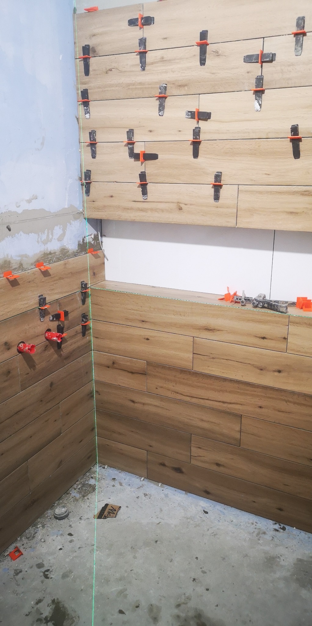 Układanie płytek imitujących drewno na ścianie w łazience, widoczne pomarańczowe klipsy poziomujące i pionowa linia lasera, ściana częściowo wykończona, surowy beton na podłodze.