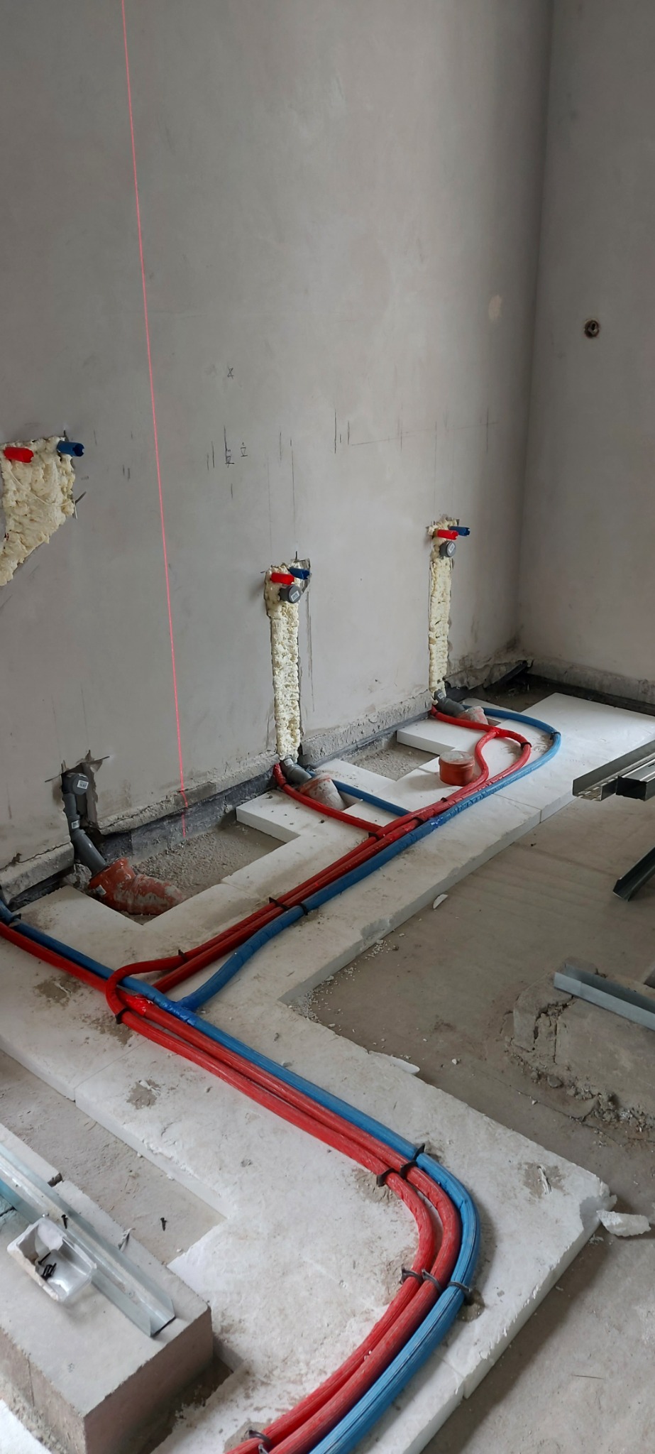 Wnętrze w trakcie prac hydraulicznych: czerwone i niebieskie rury instalacji wodnej ułożone na białych płytach izolacyjnych, pionowe rury przy ścianie zabezpieczone pianką montażową, widoczna linia...