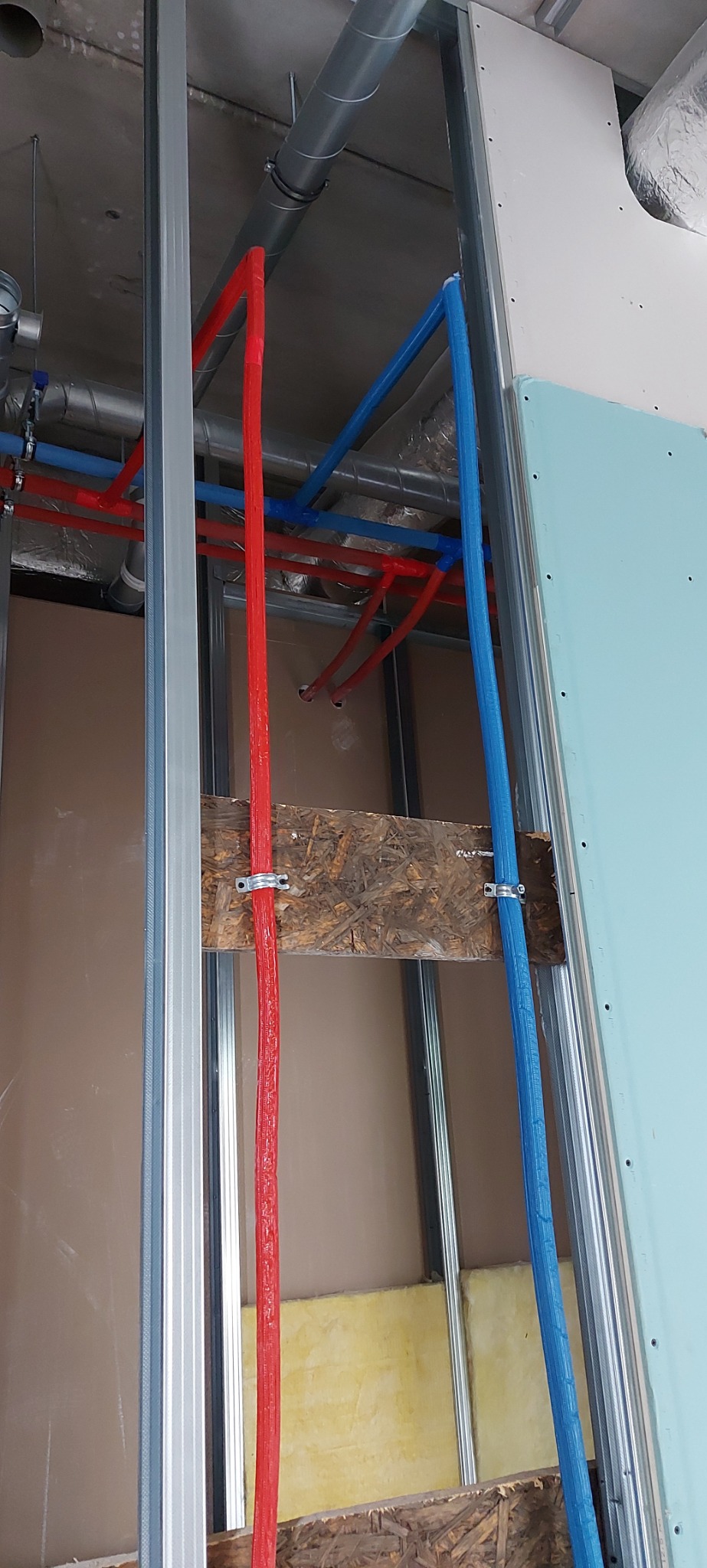 Czerwona i niebieska rura instalacji wodnej przymocowane do drewnianej belki wewnątrz ściany szkieletowej z metalowymi profilami i płytą gipsowo-kartonową.