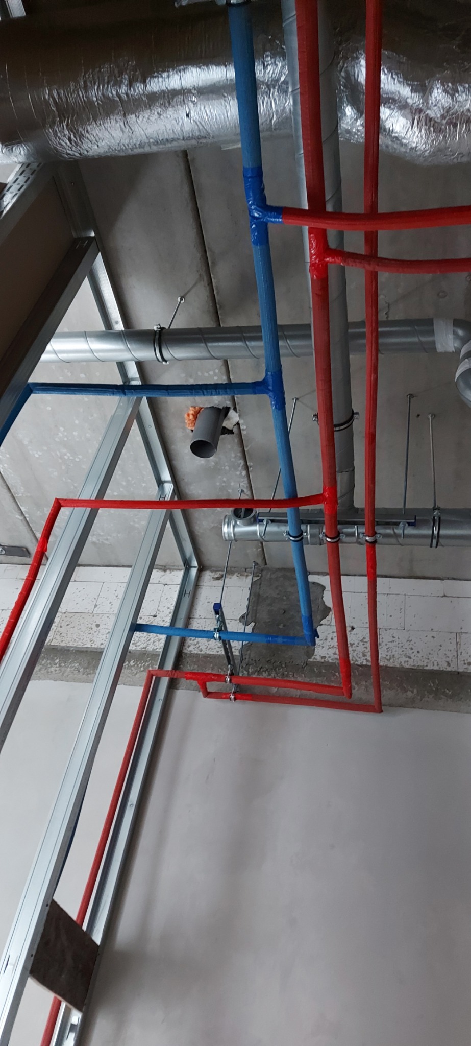 Instalacja hydrauliczna i grzewcza w trakcie montażu pod sufitem, rury w kolorach czerwonym i niebieskim, widoczne elementy konstrukcji sufitu podwieszanego i wentylacji.