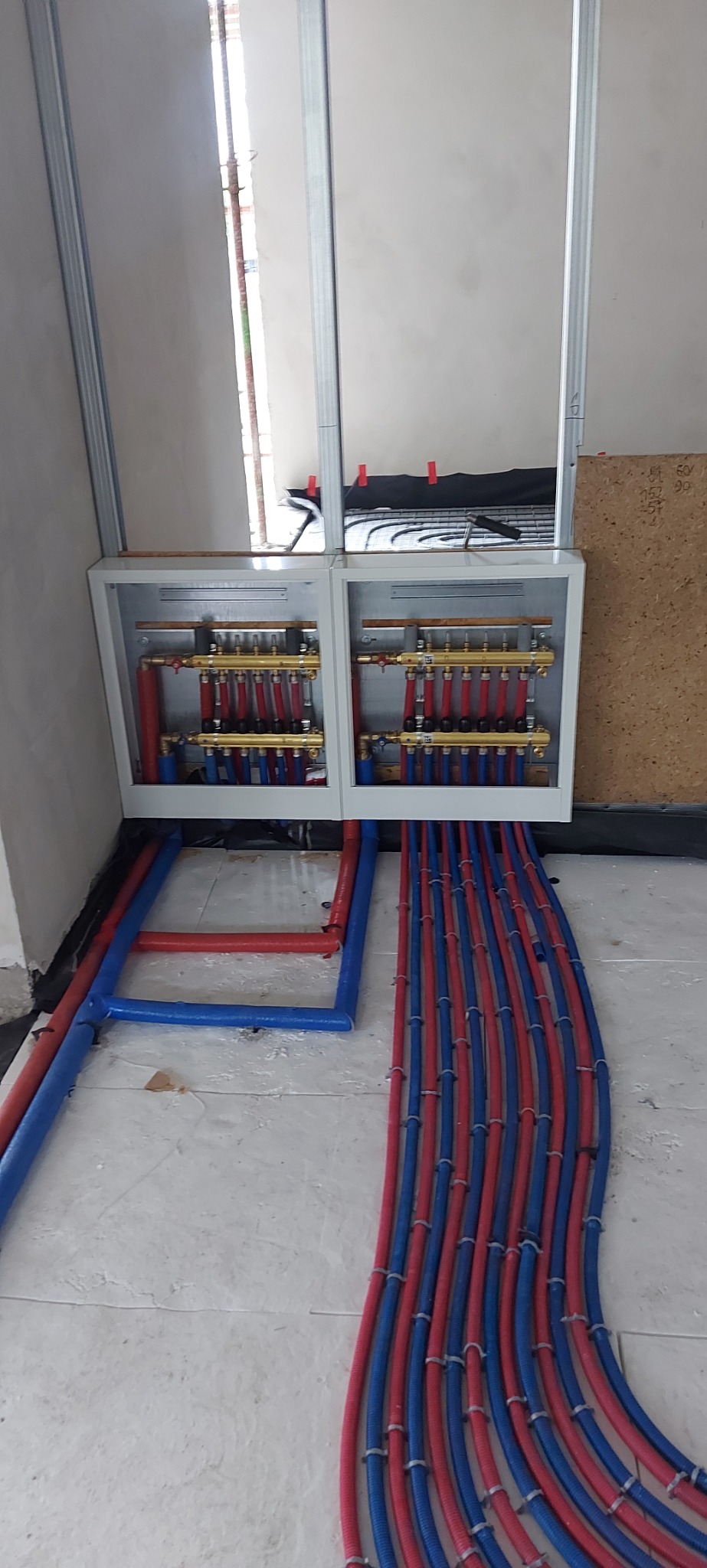Rozdzielacz ogrzewania podłogowego w szafce, z licznymi niebieskimi i czerwonymi rurami prowadzącymi do instalacji, widoczne elementy montażowe na ścianie.