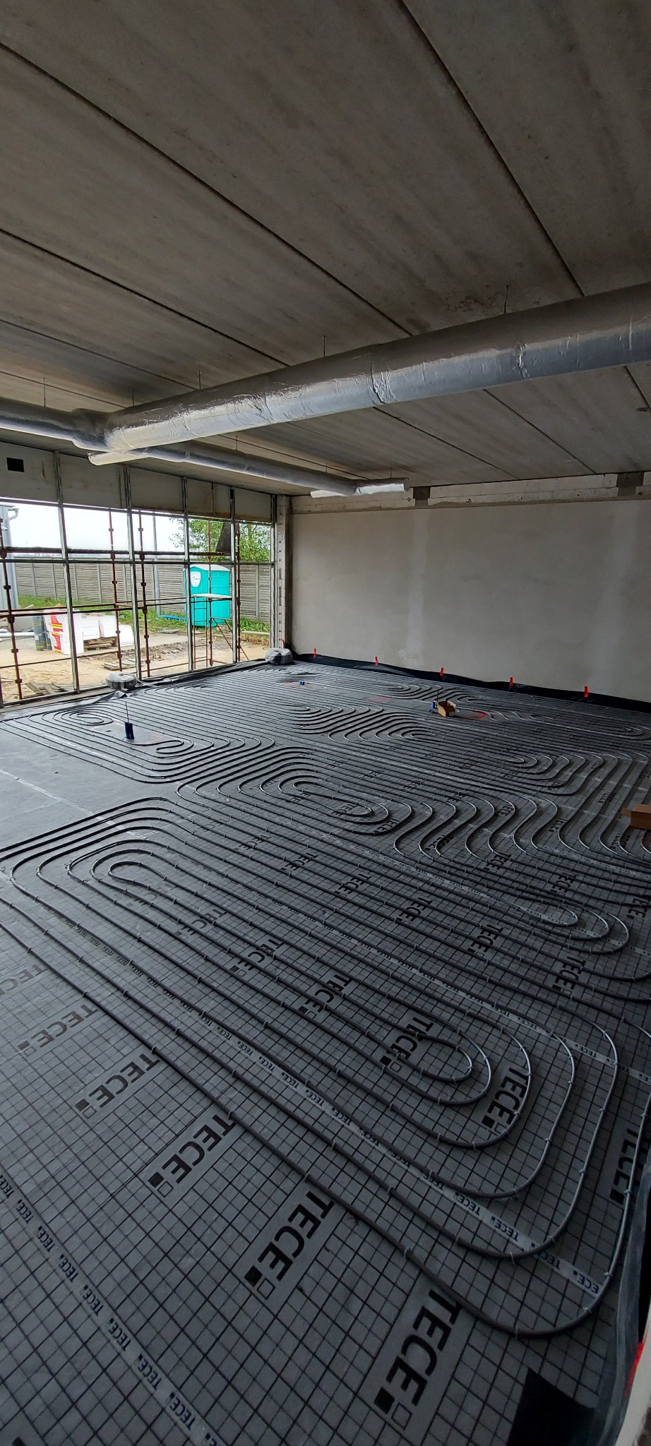 Montaż ogrzewania podłogowego w surowym wnętrzu, widoczne rury ułożone w pętle na izolacji z nadrukiem TECE, surowy betonowy sufit z instalacją wentylacyjną.