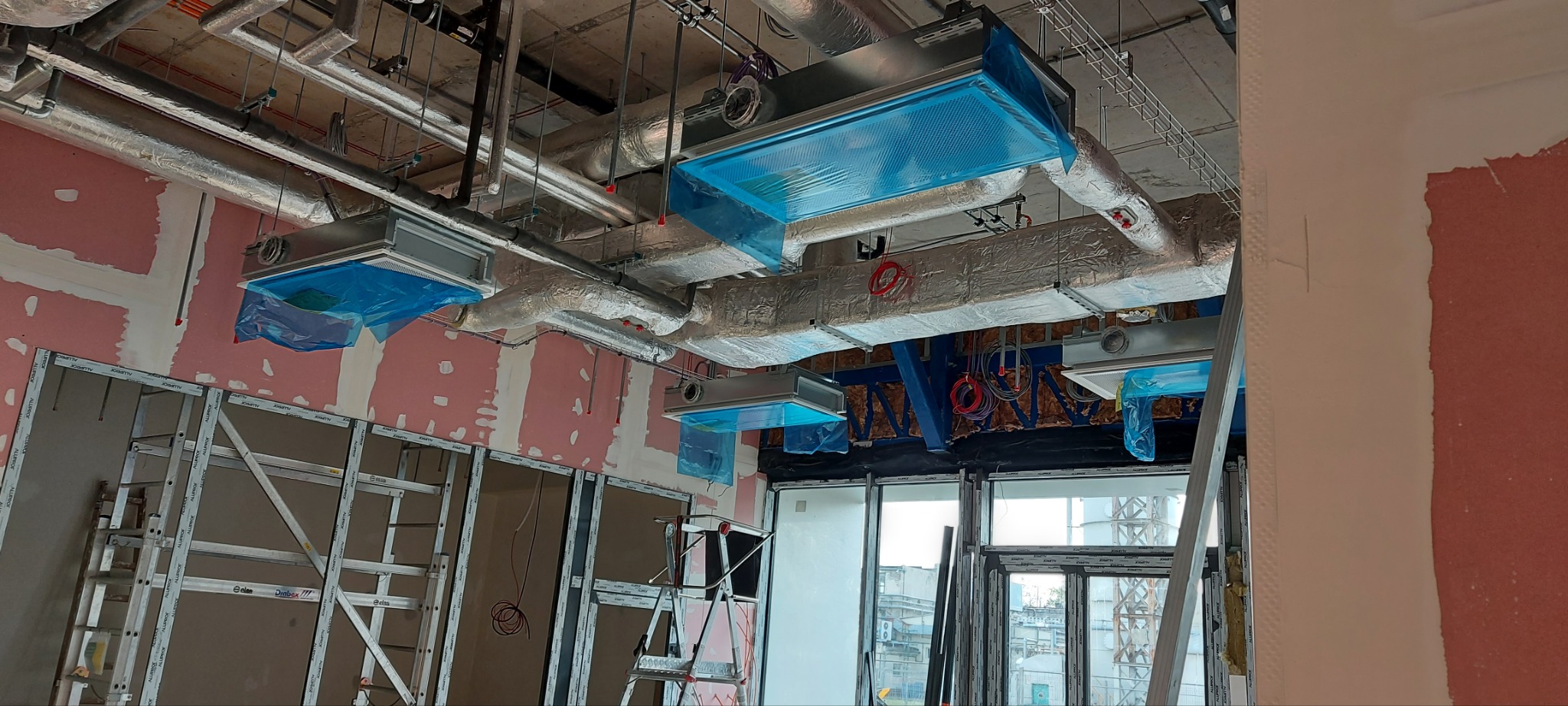 Montaż systemów wentylacyjnych pod sufitem w surowym wnętrzu, widoczne kanały wentylacyjne owinięte folią aluminiową, elementy konstrukcyjne i drabiny.