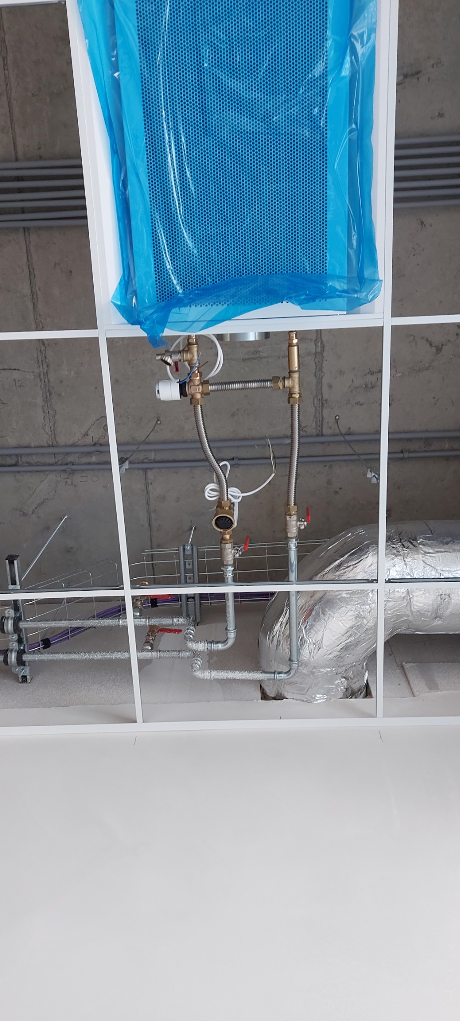 Widok instalacji grzewczej pod sufitem podwieszanym, z grzejnikiem w niebieskiej folii ochronnej, rurami miedzianymi i aluminiową izolacją termiczną.