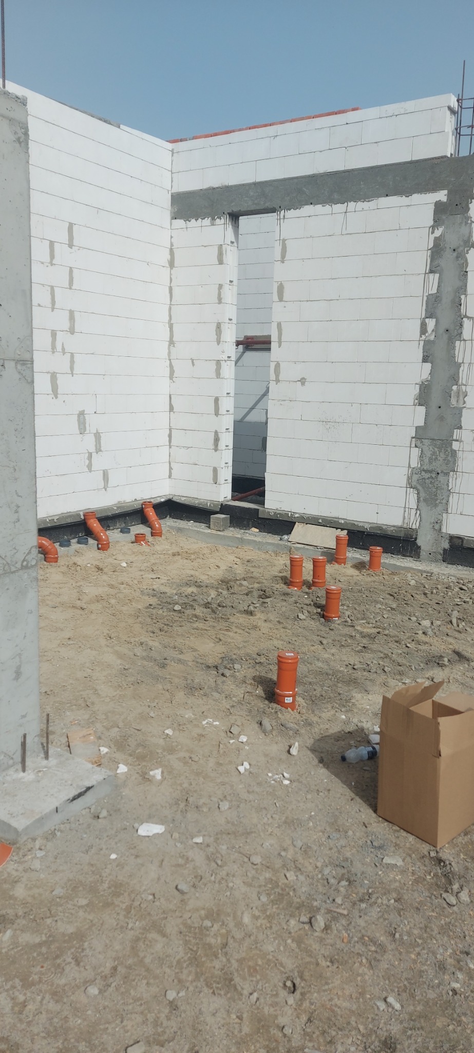 Wnętrze budynku w trakcie budowy z widocznymi, pomarańczowymi rurami instalacji kanalizacyjnej w podłożu i przy ścianach z betonu komórkowego.