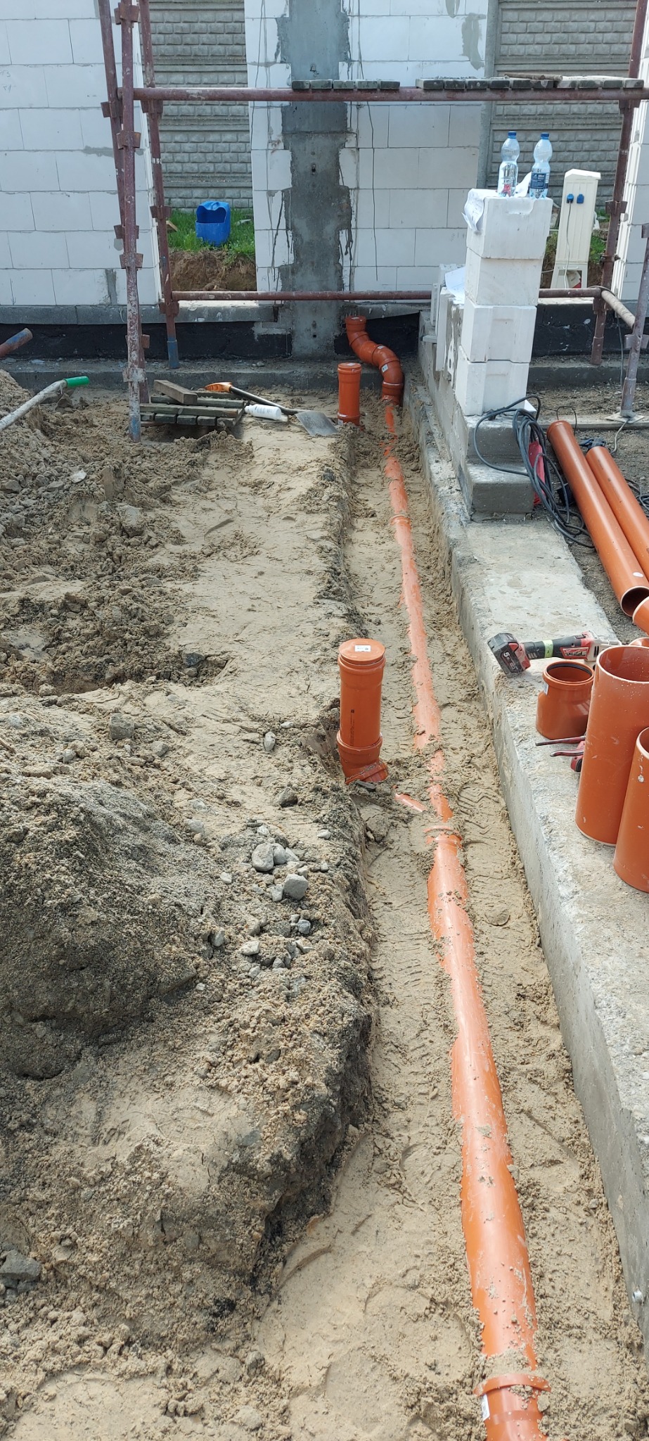 Pomarańczowe rury kanalizacyjne ułożone w wykopie ziemnym wzdłuż fundamentów budynku w trakcie budowy, widoczne narzędzia i materiały budowlane.