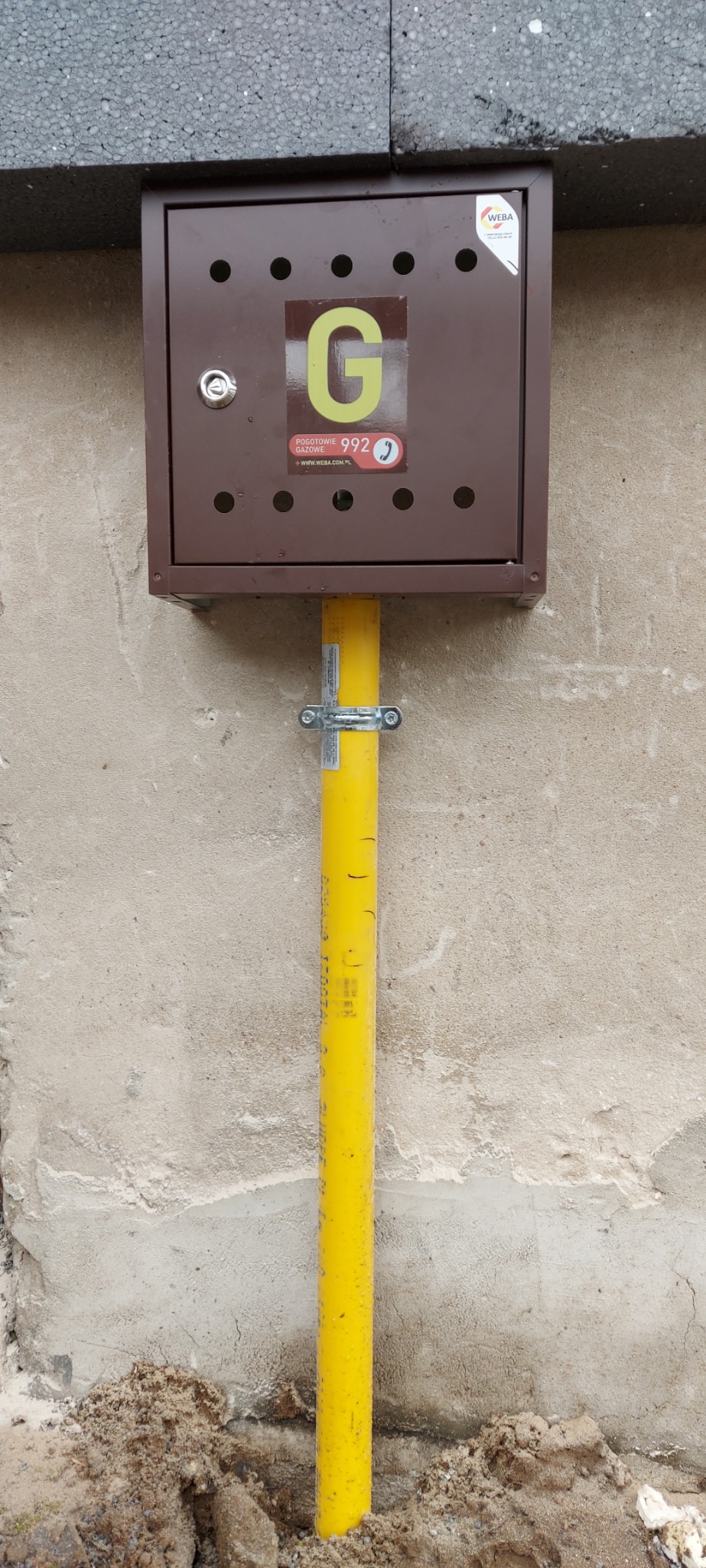 Brązowa skrzynka gazowa z żółtą rurą przymocowana do szarej ściany, widoczna ziemia u podstawy, logo WEBA na skrzynce.