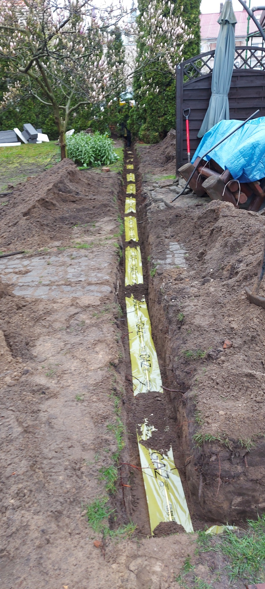 Wąski wykop w ziemi z żółtą folią ostrzegawczą z napisem 'GAZ', widok z góry, w tle ogród z drzewami i krzewami.