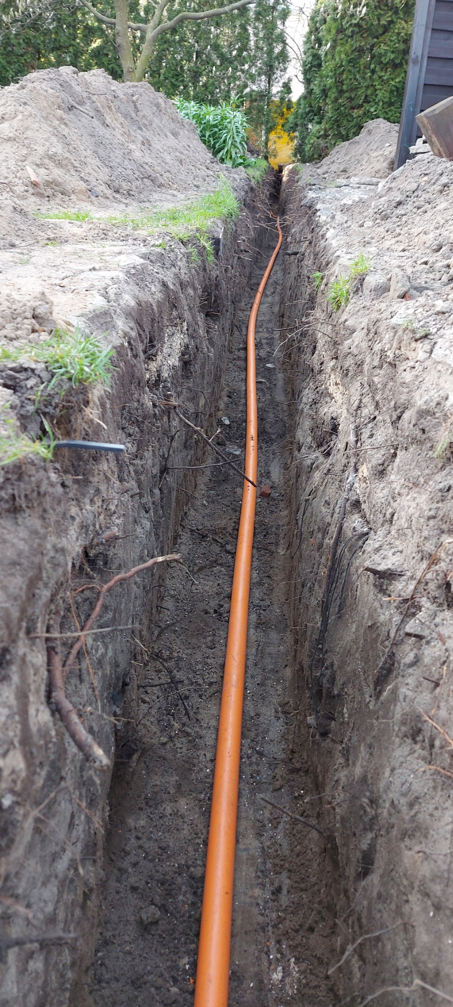 Pomarańczowa rura w wąskim wykopie w ziemi, widok z góry, z fragmentami zieleni na krawędziach wykopu.