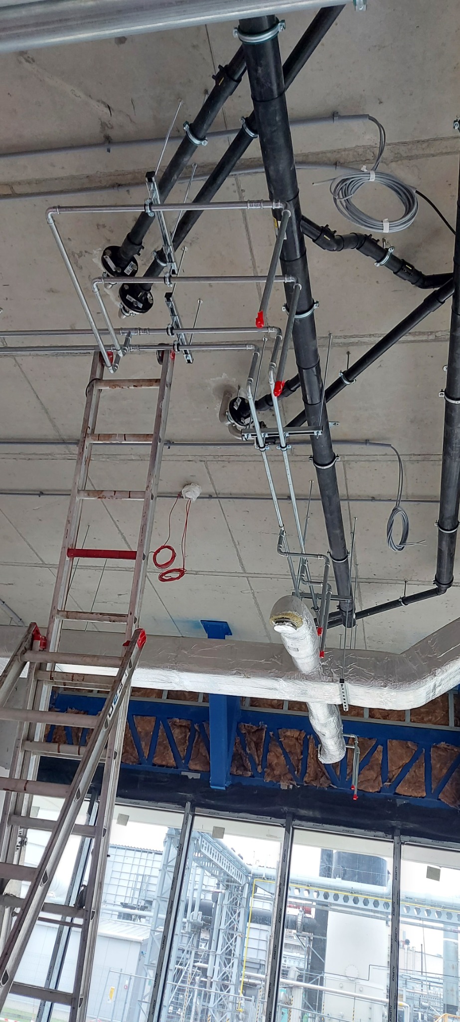 Widok z dołu na instalacje pod sufitem w trakcie montażu, widoczna drabina aluminiowa oparta o okno, rury czarne i srebrne mocowane do sufitu, przewody elektryczne, instalacja przeciwpożarowa...