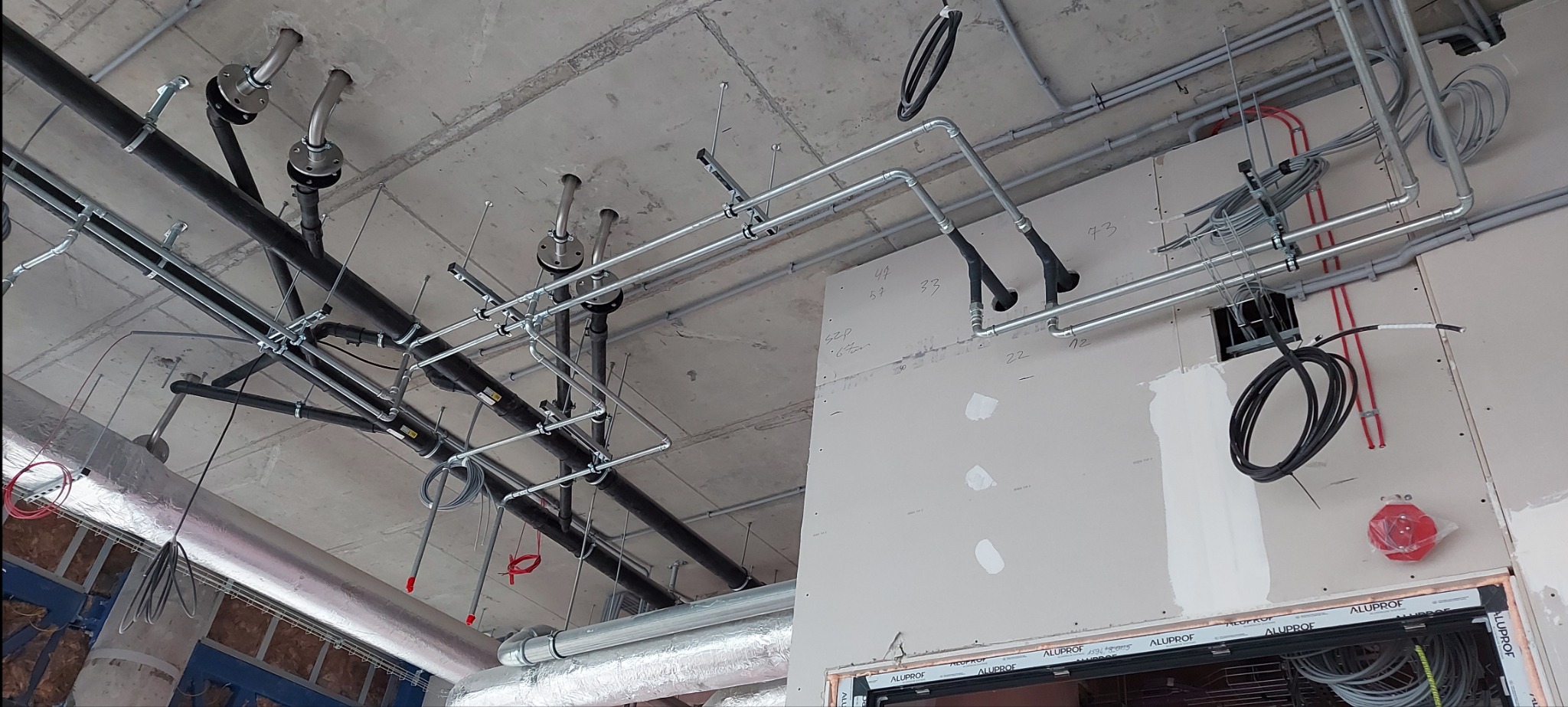 Widok sufitu w trakcie montażu instalacji: rury stalowe, czarne rury kanalizacyjne, przewody elektryczne, widoczna izolacja termiczna rur, ślady po wierceniach w płycie gipsowej.