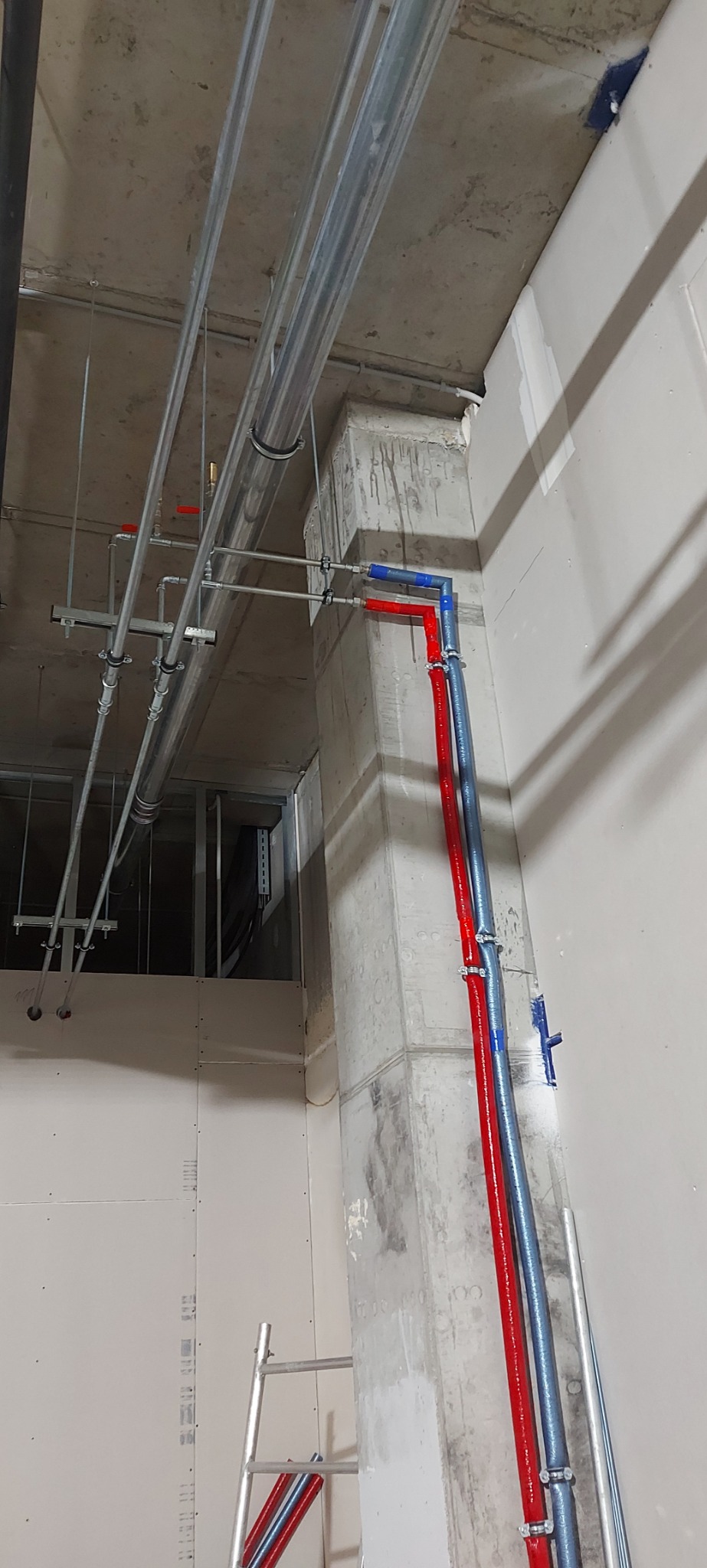 Wnętrze w trakcie prac wykończeniowych z widocznymi rurami instalacji w kolorach czerwonym i niebieskim, przymocowanymi do betonowego filara. Nad nimi widoczne metalowe rury podwieszone do sufitu...