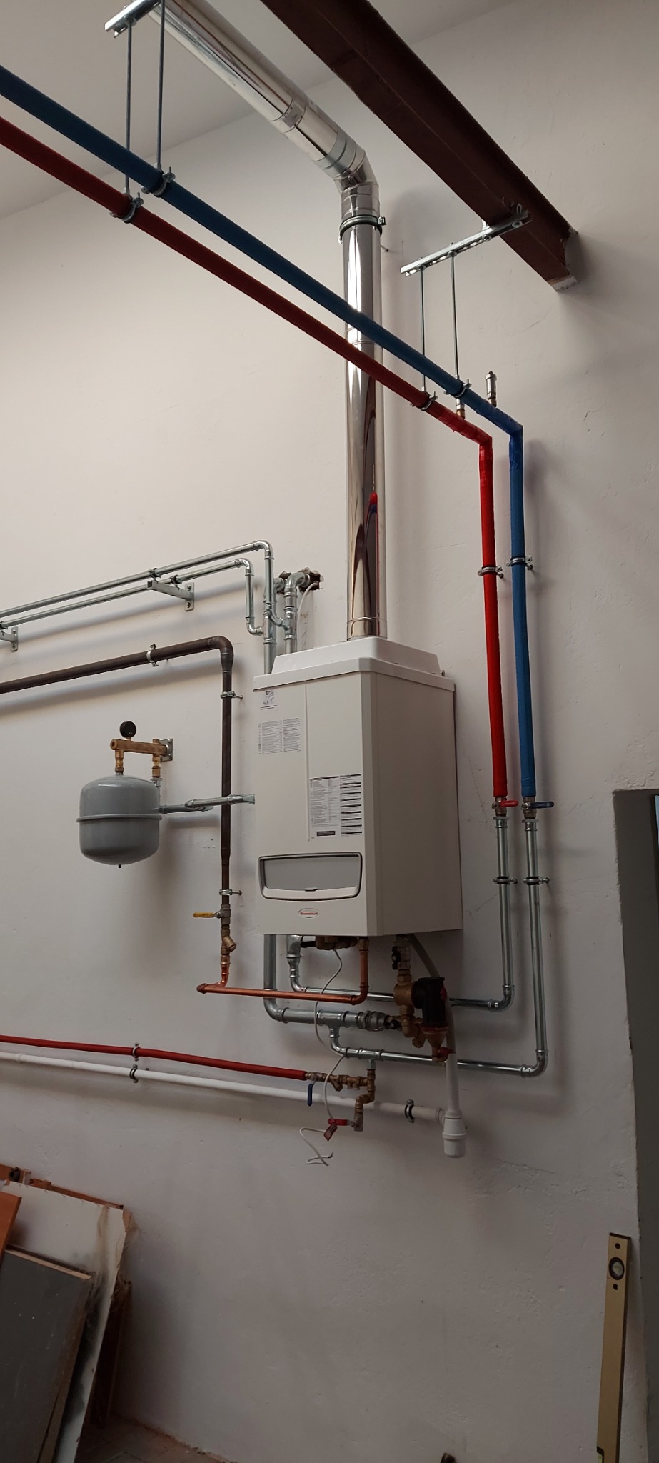 Nowoczesny, biały piec gazowy z podłączonymi rurami miedzianymi i stalowymi, w tym komin ze stali nierdzewnej, oraz rury grzewcze w kolorze czerwonym i niebieskim, zamontowany na białej ścianie.