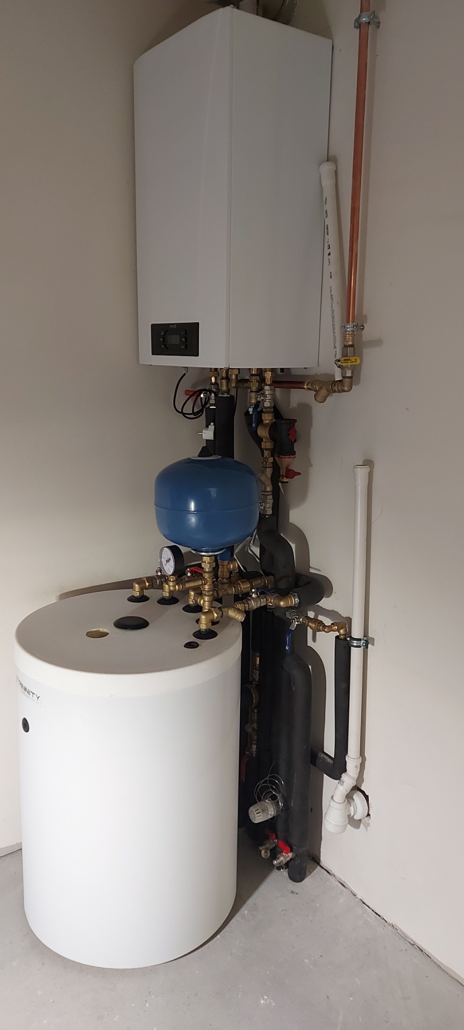 Nowoczesny, kompaktowy system grzewczy z białym zasobnikiem ciepłej wody i wiszącym kotłem, z licznymi rurami miedzianymi i czarnymi izolowanymi przewodami, zainstalowany w rogu pomieszczenia.